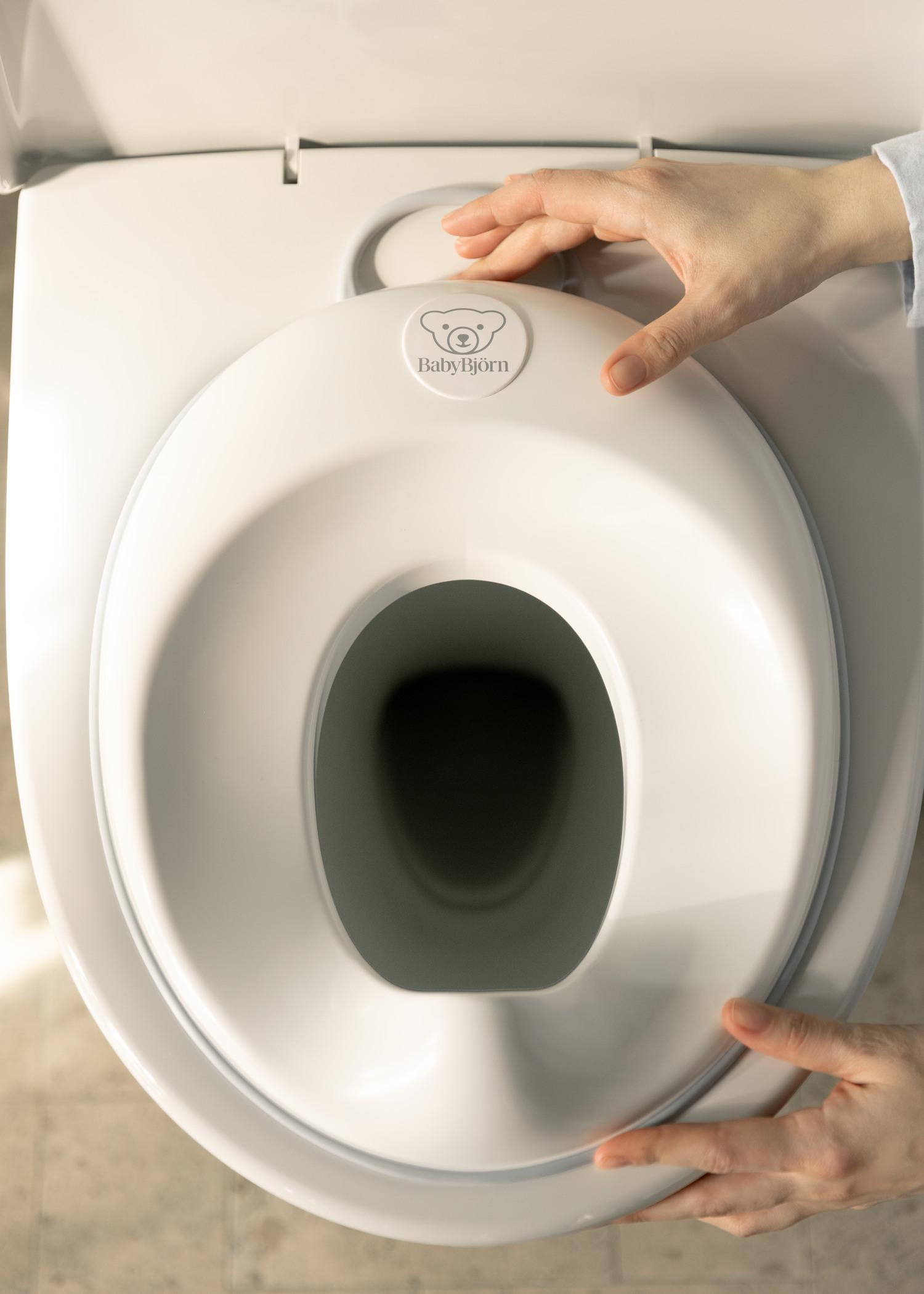 Hands holding a white BabyBjörn potty seat placed on a toilet, viewed from above.