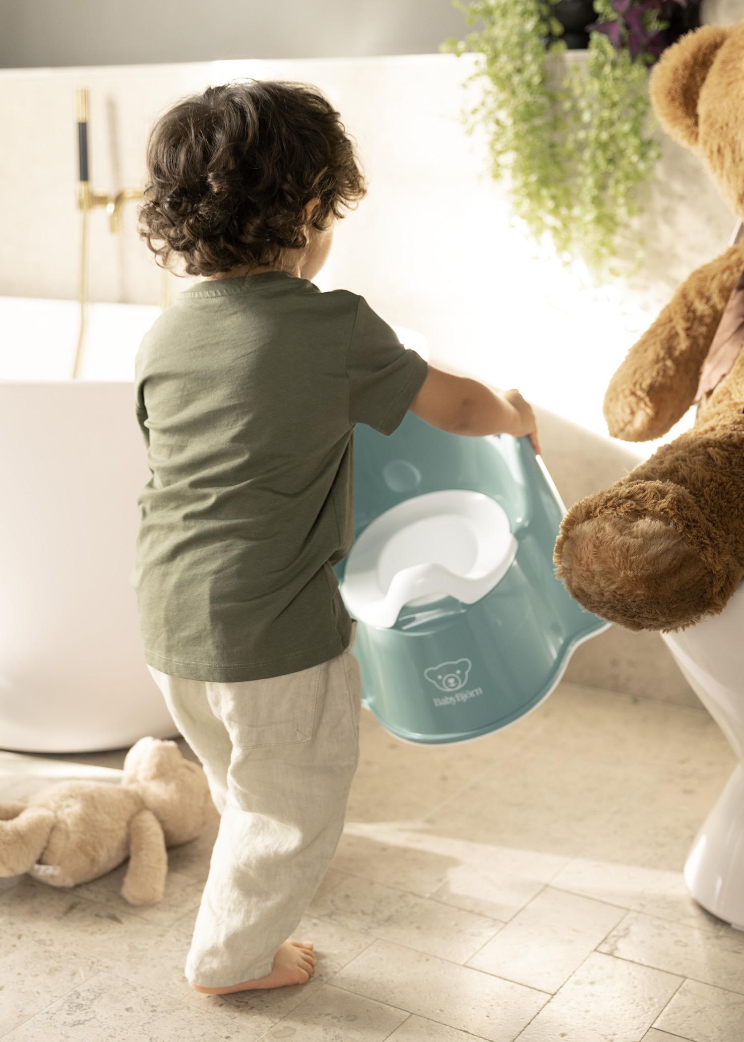 Child holding a blue potty chair in a bathroom, with a large teddy bear nearby and a plush toy on the floor.