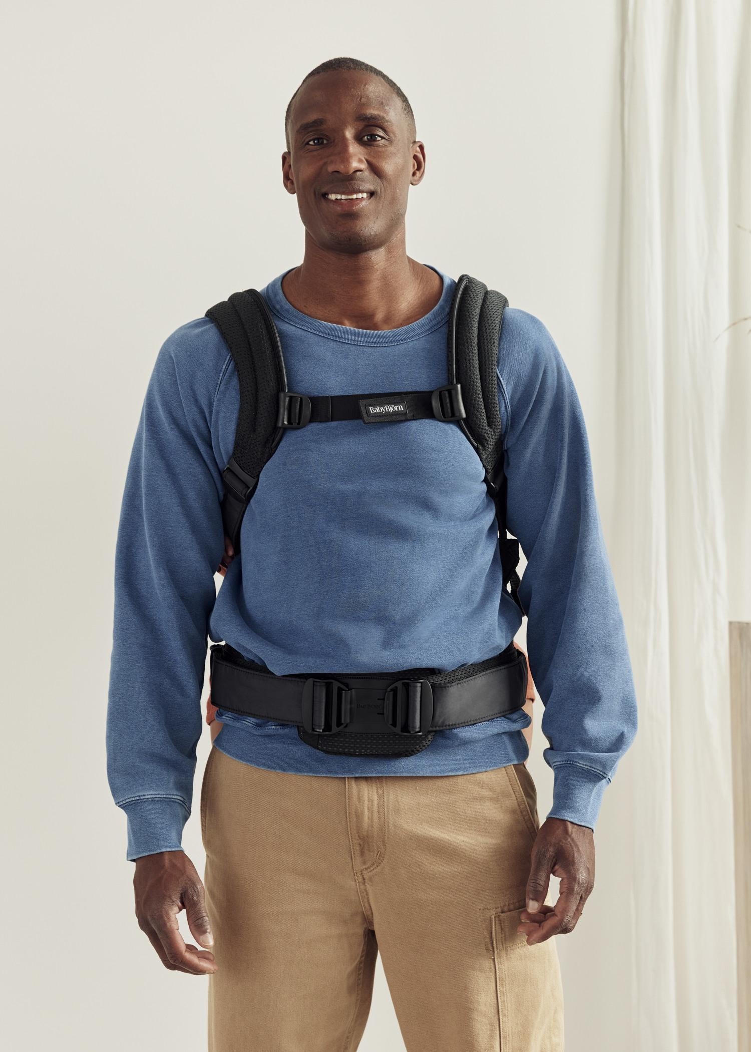 Man in blue sweatshirt wearing a black baby carrier over his shoulders and waist, standing in a brightly lit room.