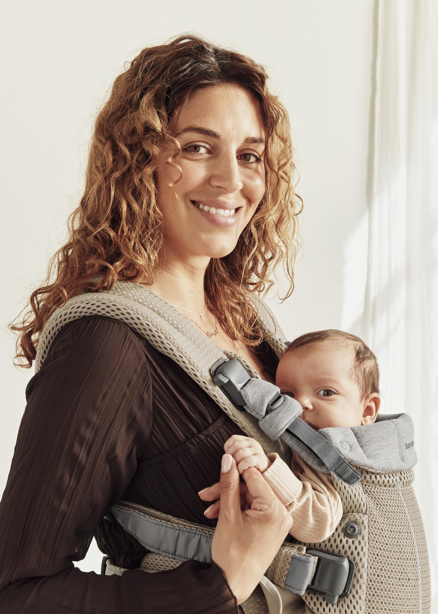 Woman with curly hair smiles while carrying a baby in a gray beige baby carrier.