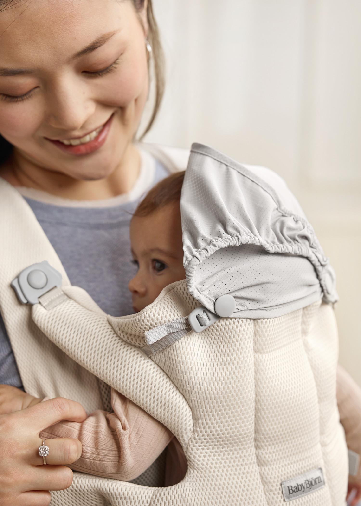 A smiling woman holds a baby in a beige BabyBjörn carrier. The baby, wearing a hood, is nestled comfortably against her chest.