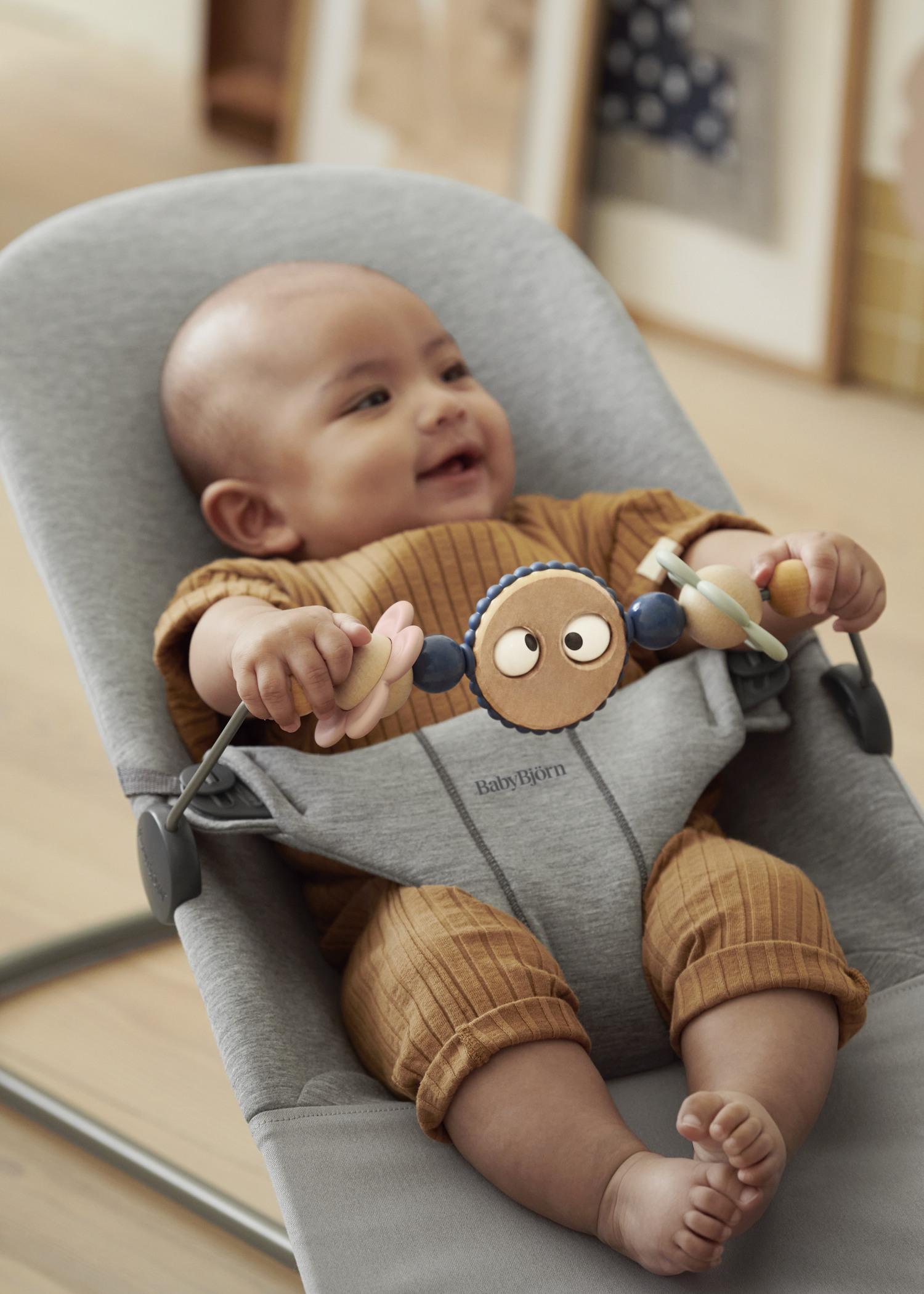 A smiling baby in a brown outfit sits in a gray bouncer holding a toy with colorful beads and faces.