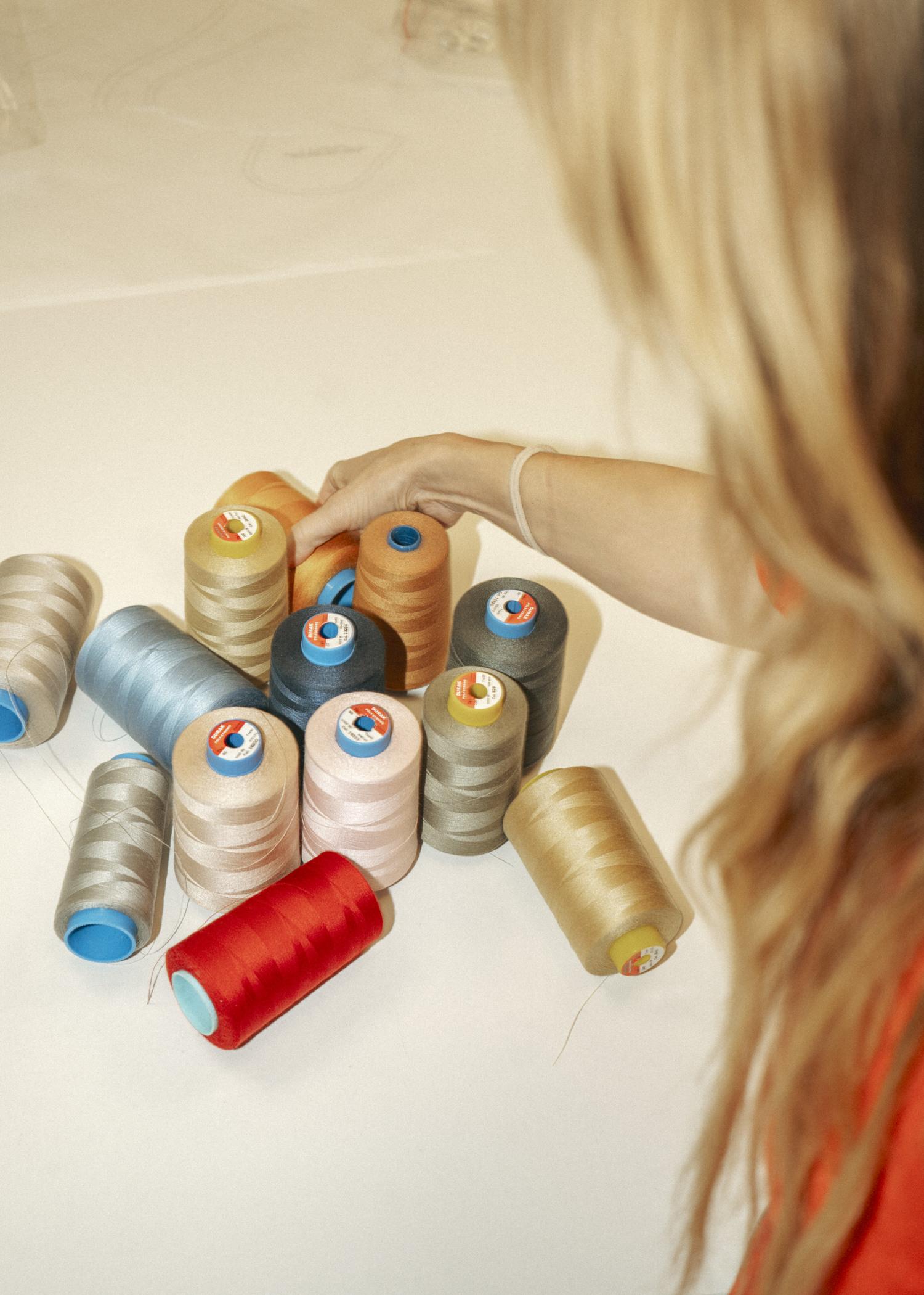 A person with long hair selects from an array of colorful thread spools on a table, including red, blue, silver, and brown shades.