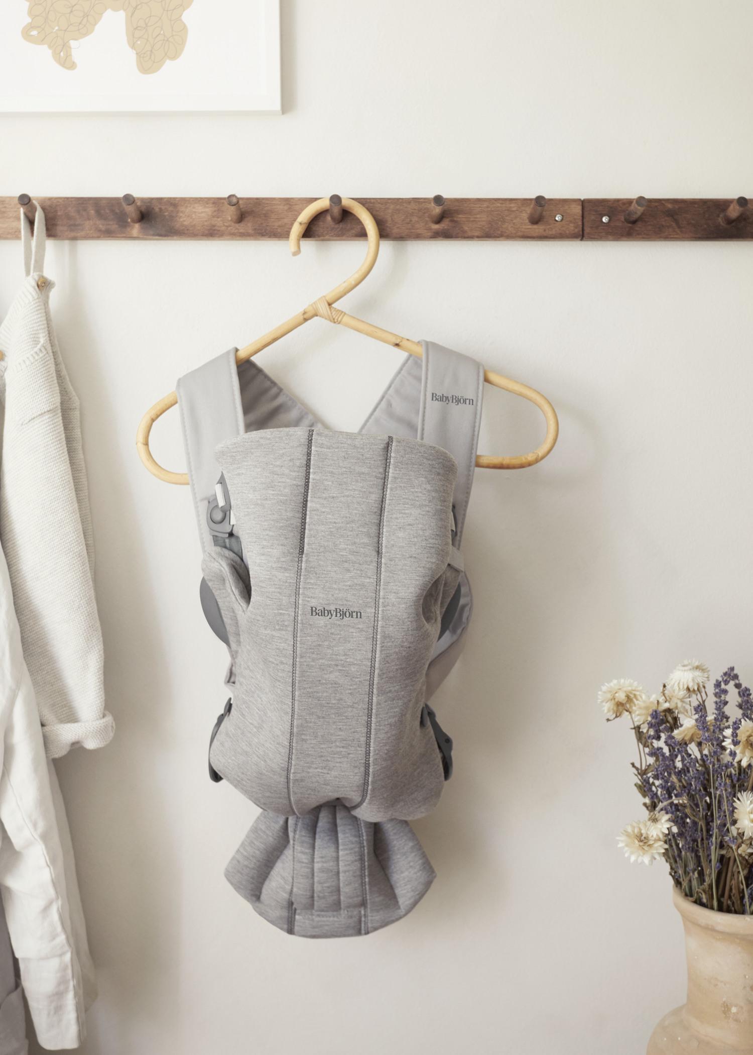 Gray baby carrier hanging on a wooden peg, with a beige sweater beside it and dried flowers in a vase to the right.