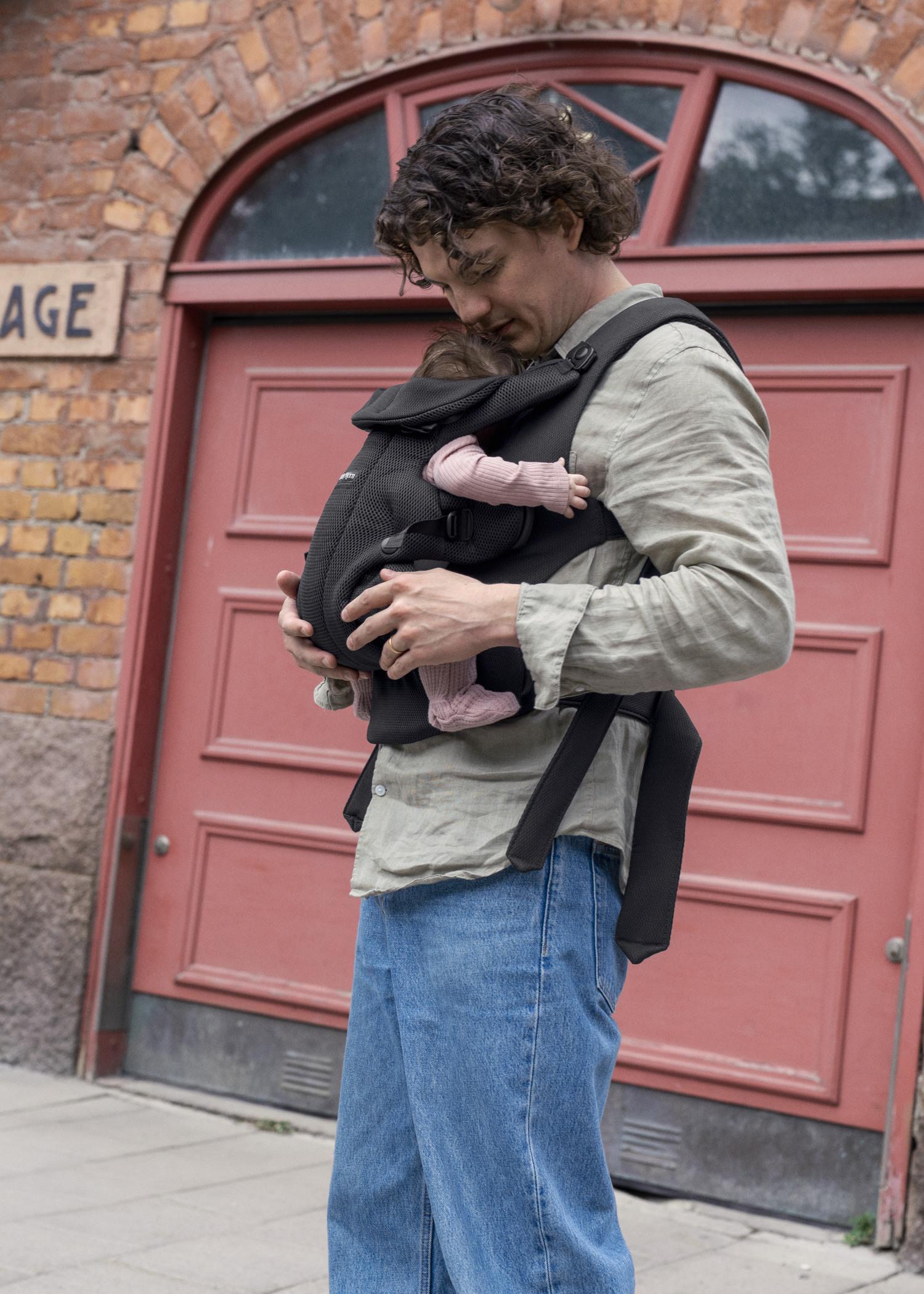 A man stands in front of a red brick building, gently holding a baby in a black carrier strapped to his chest.