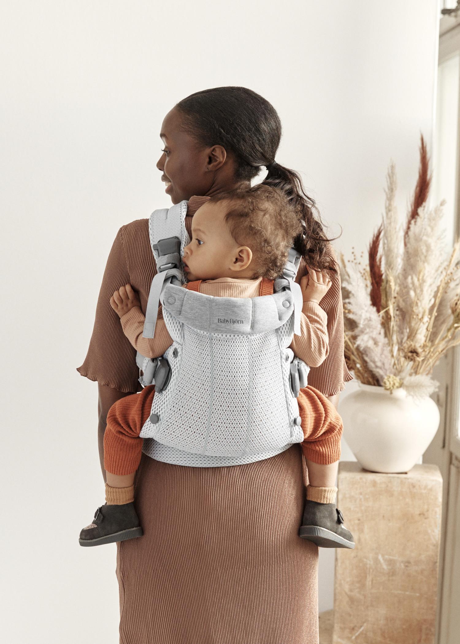 A woman wearing a brown dress carries a baby on her back in a light gray baby carrier. The baby wears a brown outfit and looks to the side.