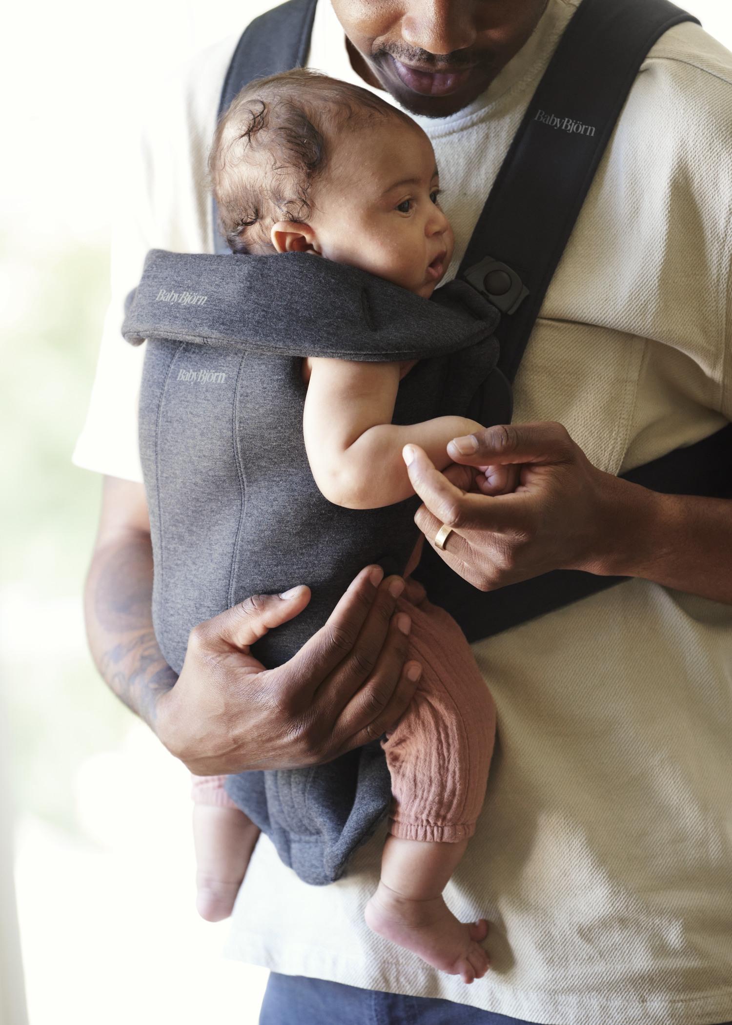 A man holding a baby in a gray carrier, gently touching the baby's hand. The baby is wearing a pink outfit.