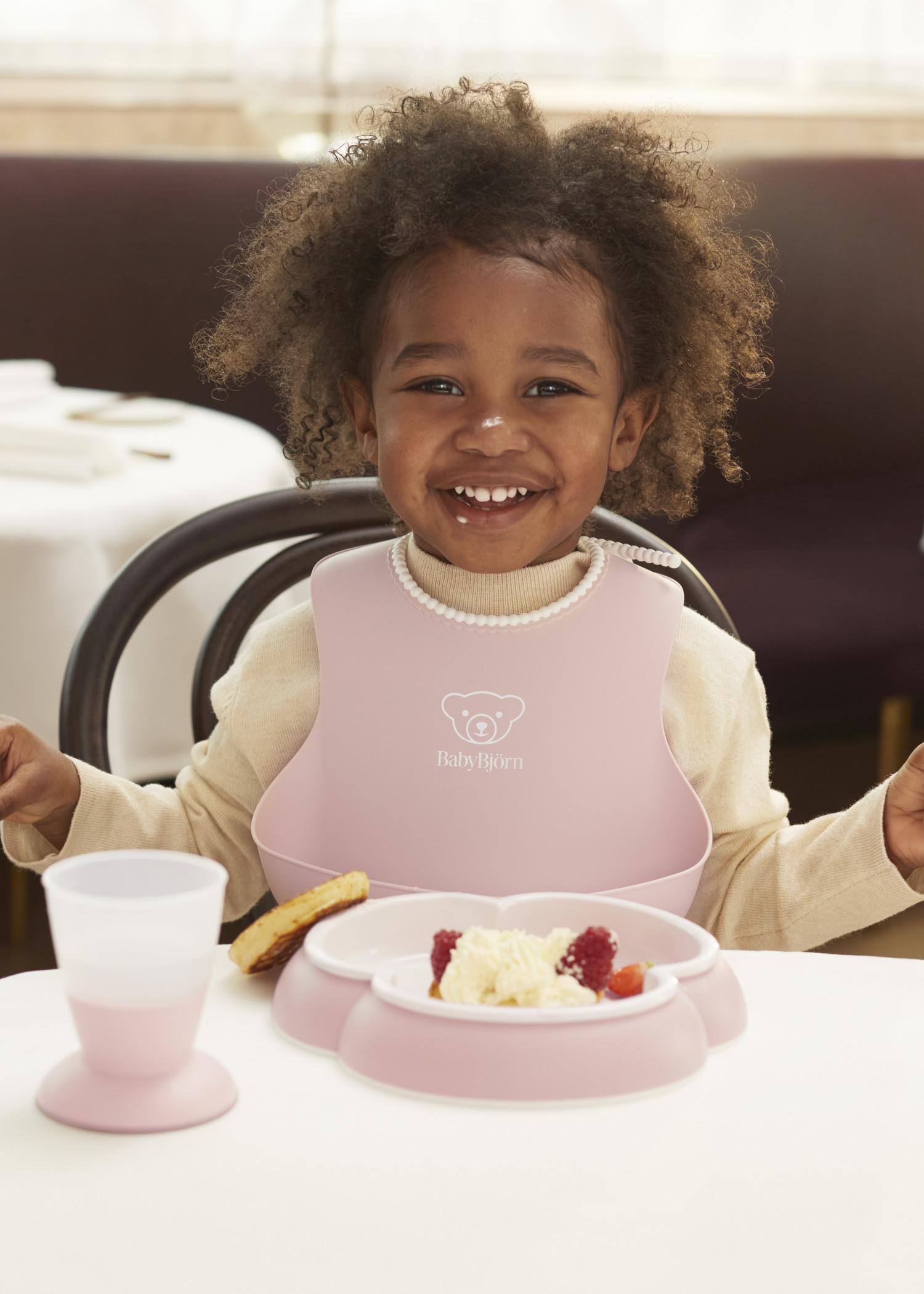 Smiling child with curly hair wearing a pink bib, sitting at a table with a pink dish of food and a cup.