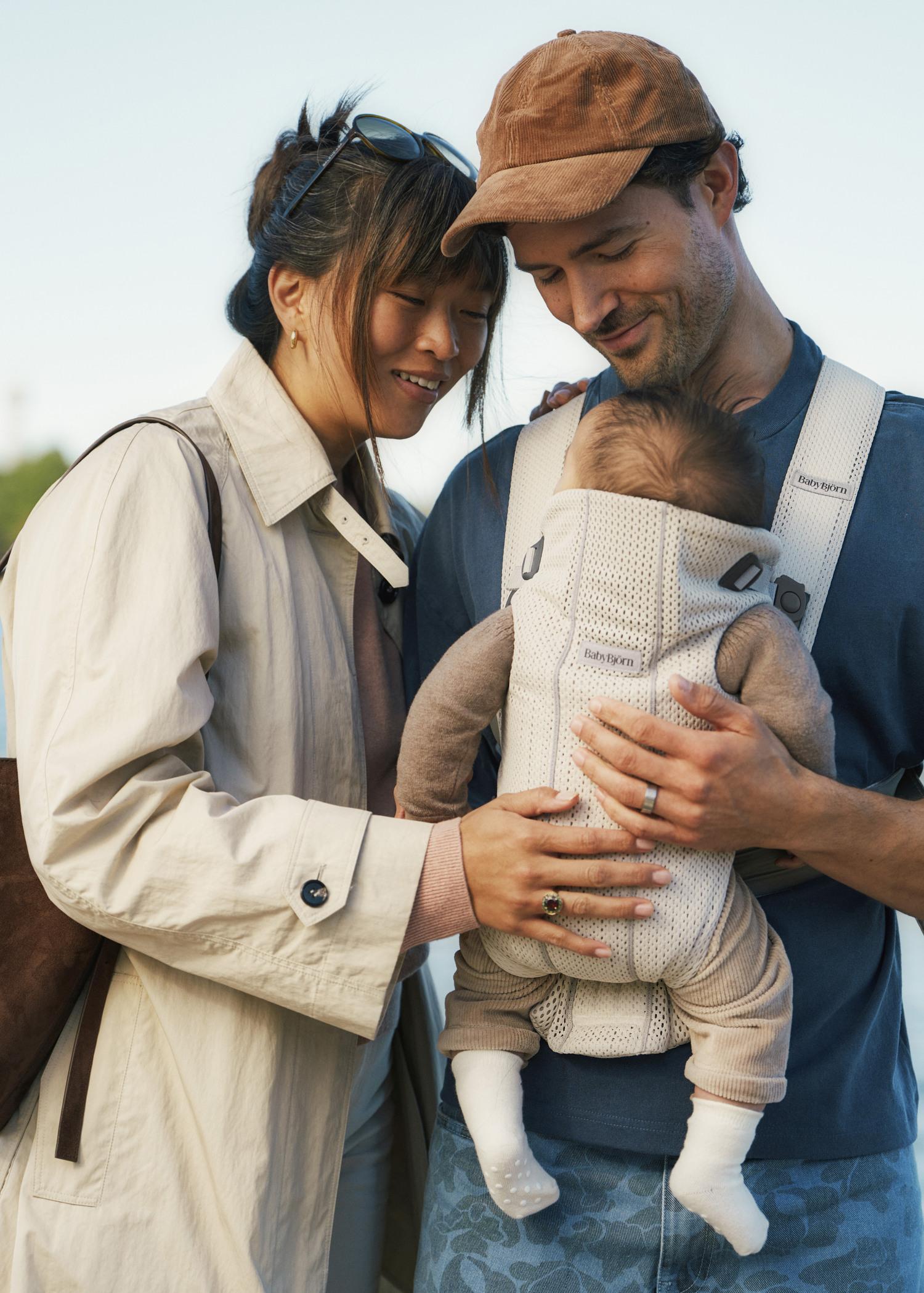 A couple smiles at their baby in a carrier. The mother wears a light coat and the father a cap, looking lovingly at the infant dressed in beige.