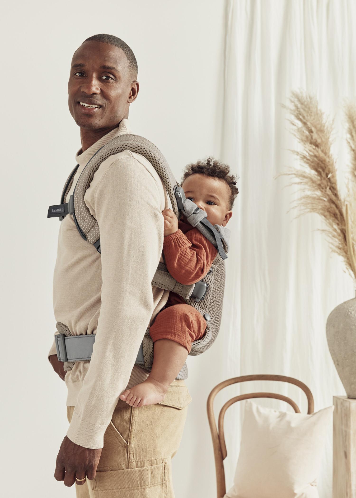 Man carrying a baby in a gray carrier, standing in a softly lit room with pampas grass and a wooden chair in the background.