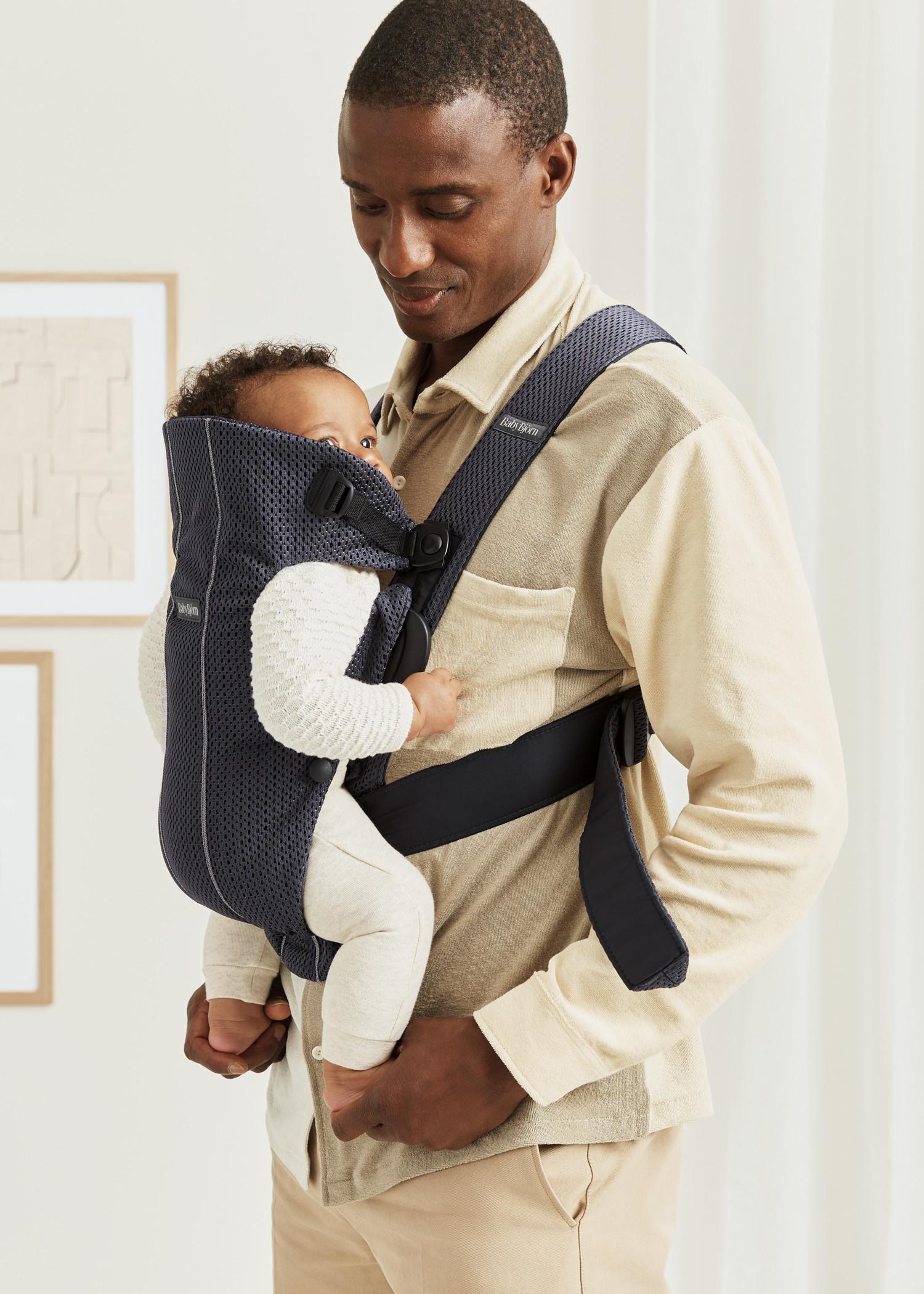 A man carrying a baby in a blue carrier, the baby is wearing a beige outfit, and they are in a softly lit indoor setting.