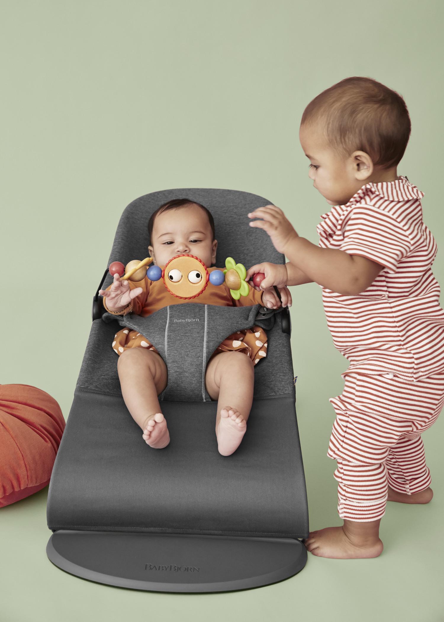 Two babies, one sitting in a baby bouncer with a toy, the other standing beside, wearing striped pajamas, on a light green background.