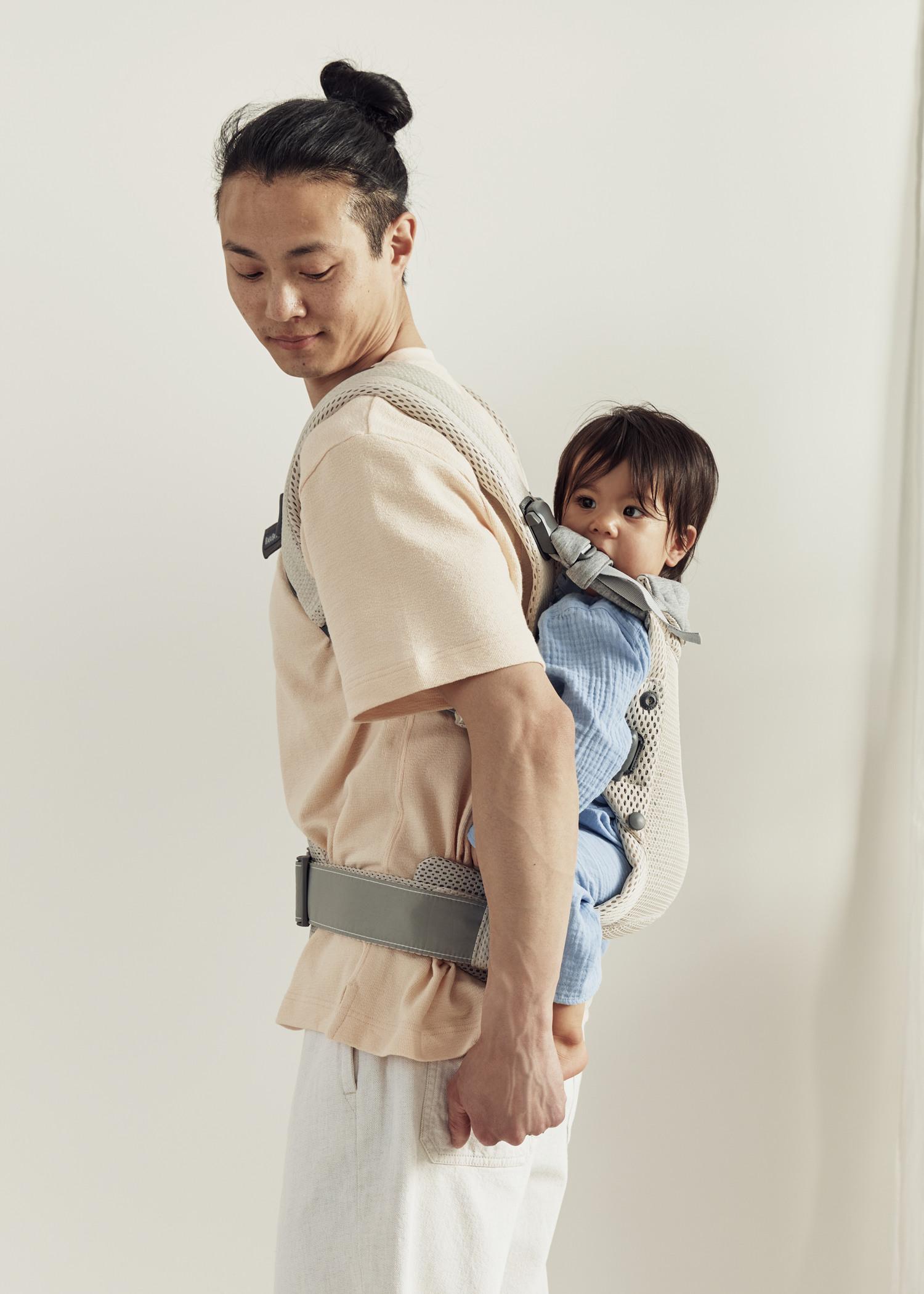 A person is wearing a light-colored outfit, carrying a baby in a blue outfit in a gray baby carrier on their back, against a plain background.