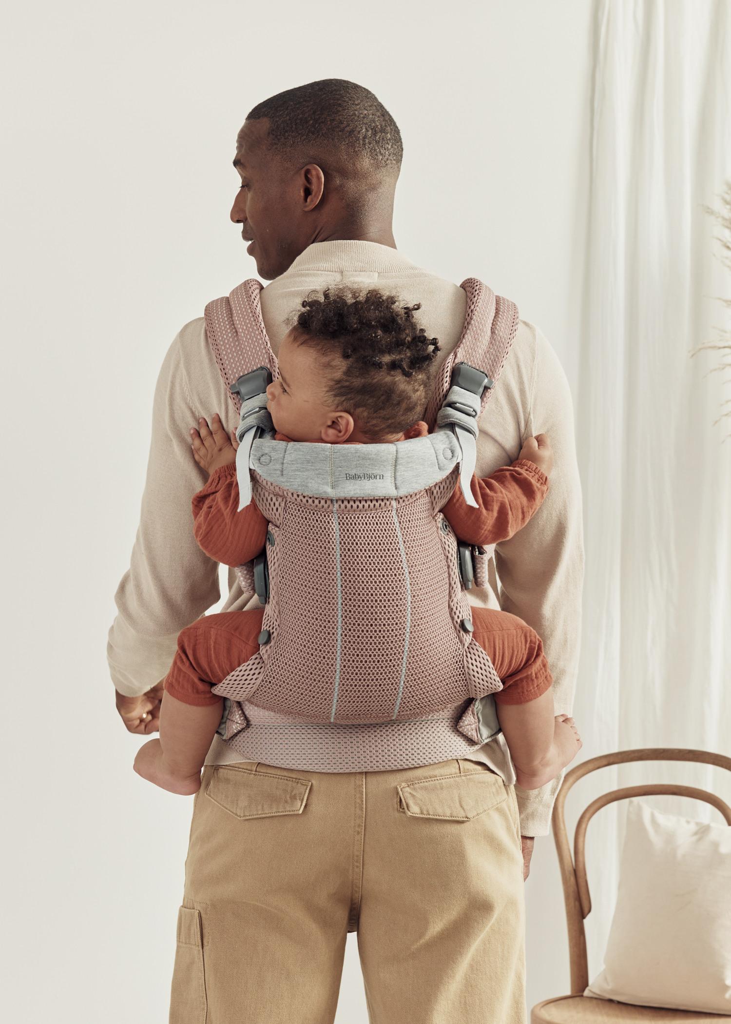 A man stands with his back to the camera, carrying a baby in a pink baby carrier. The baby is looking to the side, wearing an orange outfit.