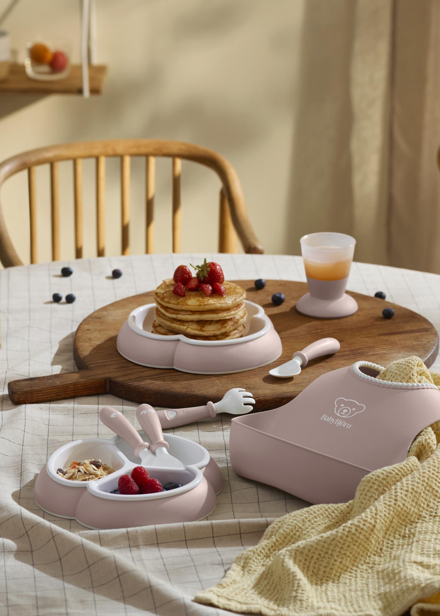 A breakfast setting with pancakes topped with strawberries, toddler utensils, a bib, and a drink on a checkered tablecloth.