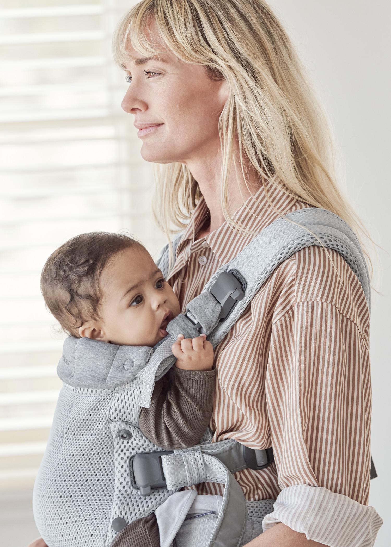 A woman with long blonde hair wearing a striped shirt carries a baby in a gray baby carrier. The baby looks to the side, gripping the carrier.