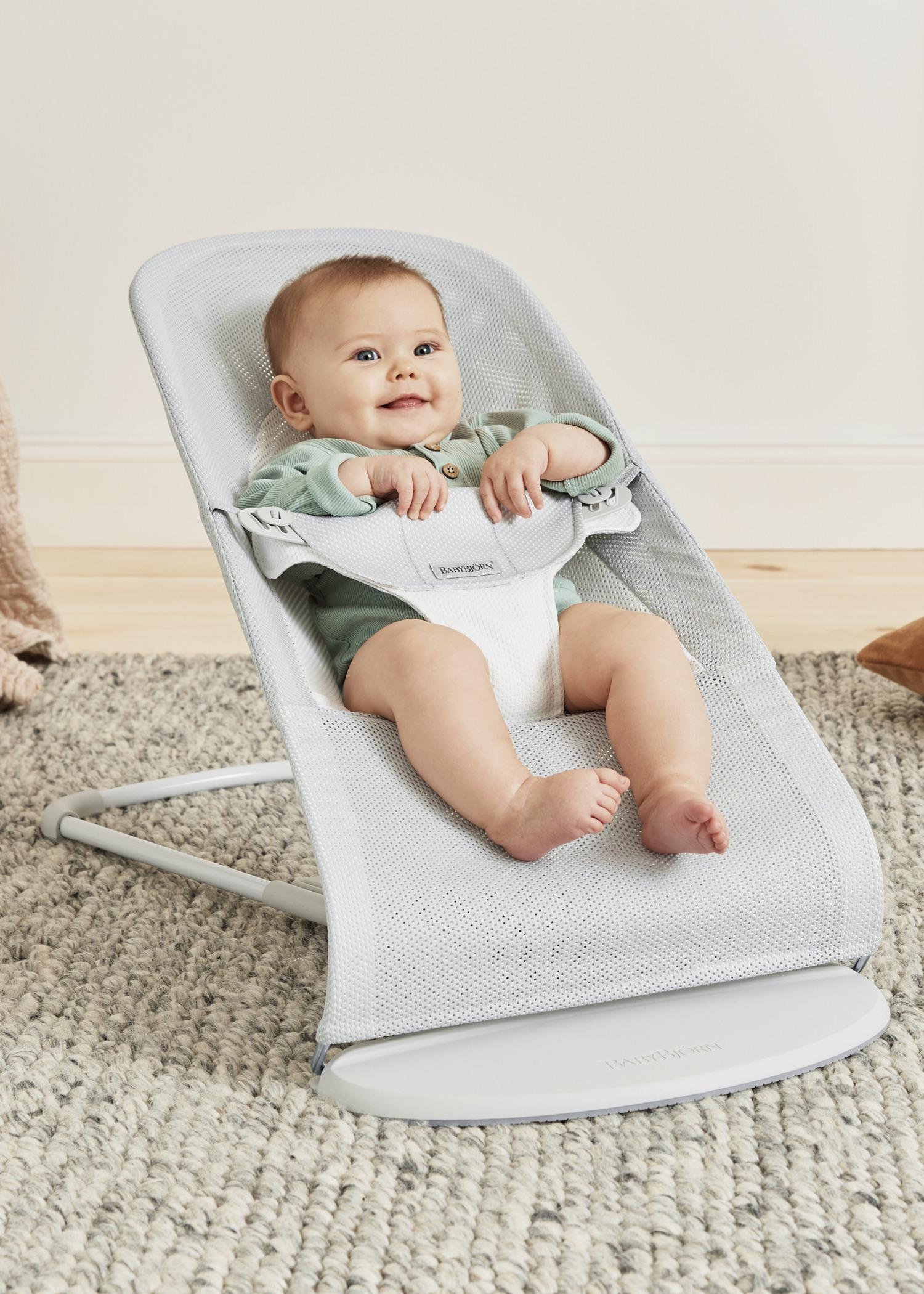 A baby in a light blue bouncer smiles while sitting on a gray carpeted floor.