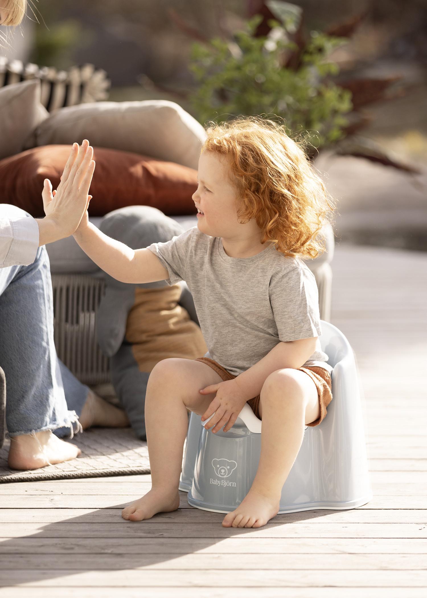 Child with curly red hair sitting on a potty, giving a high-five to an adult on a sunny patio.