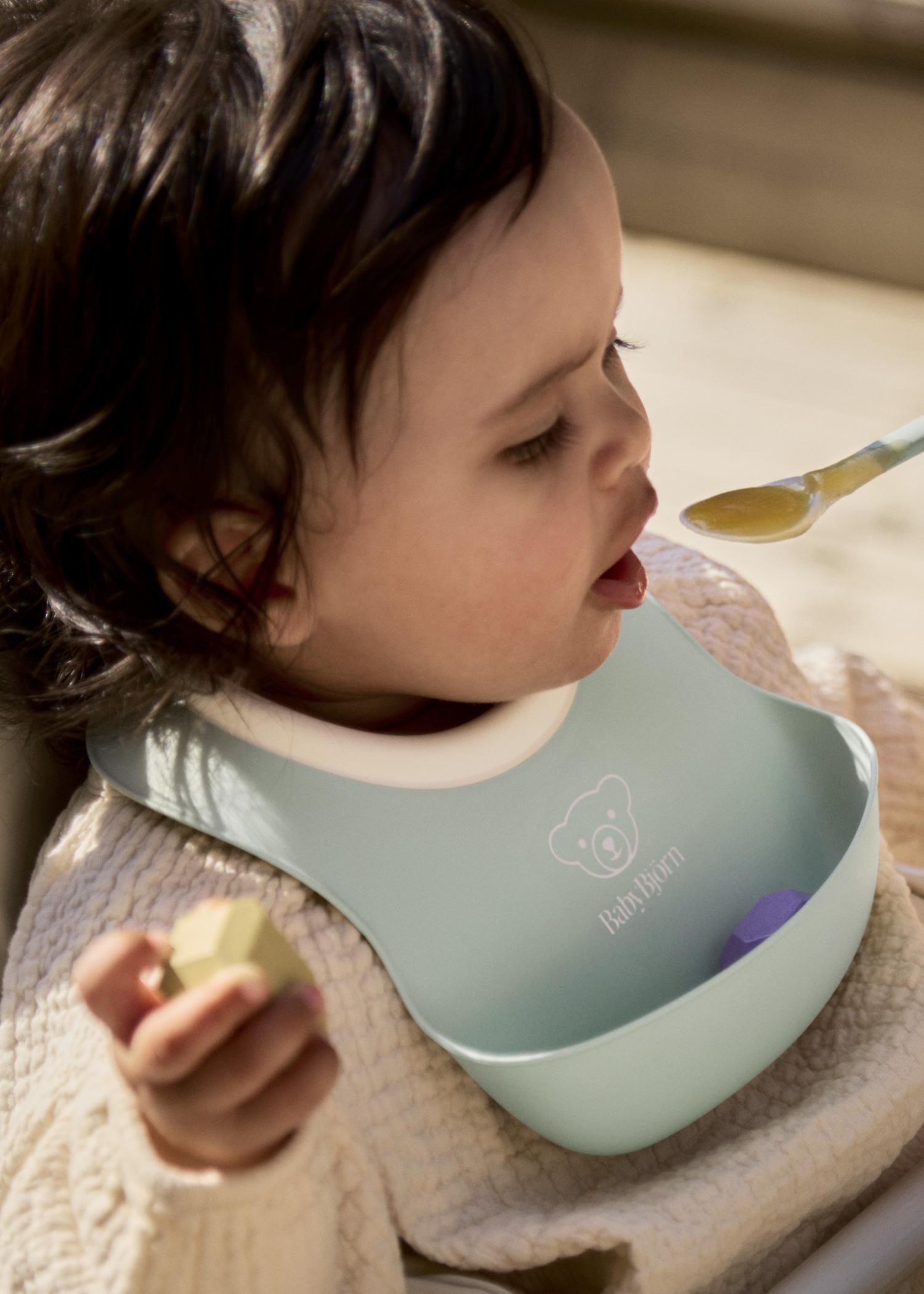 A toddler wearing a blue bib is being fed with a spoon while holding a small piece of food in one hand.