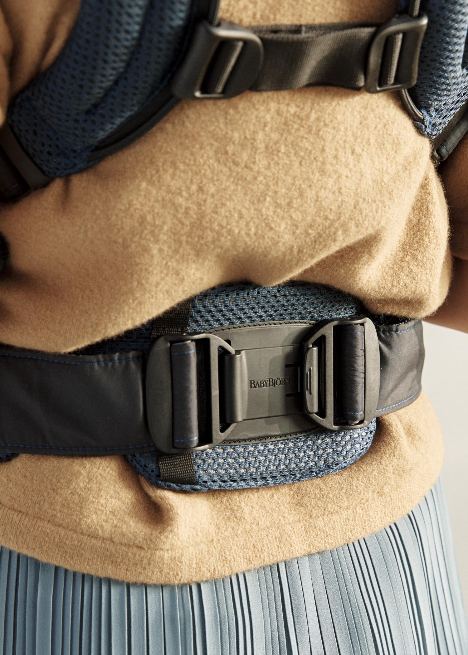 Close-up of a person wearing a baby carrier with a secure buckle, over a beige sweater and pleated blue skirt.