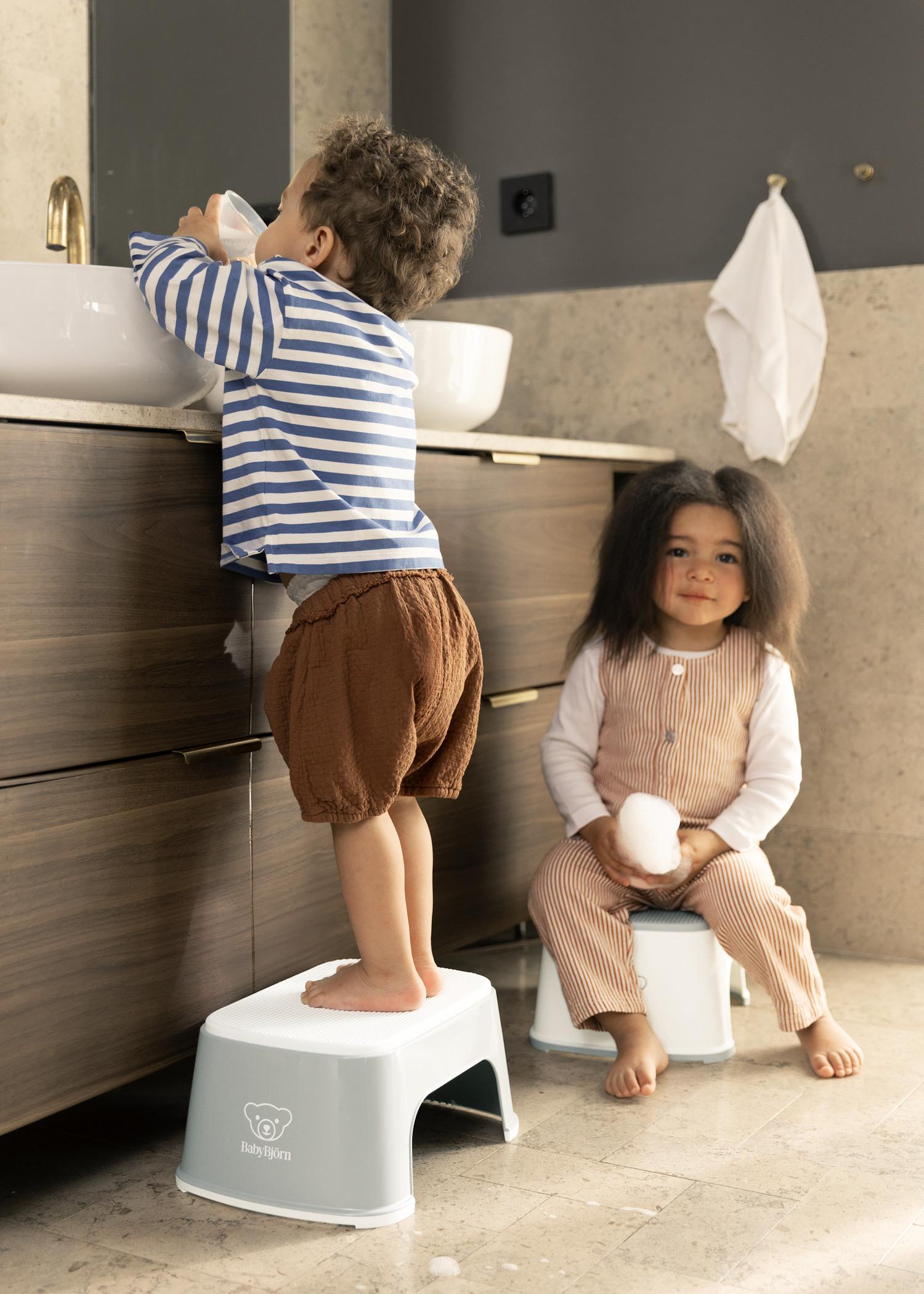Two children in a bathroom; one stands on a stool at the sink, and the other sits on a stool holding a soap bar, both in casual outfits.
