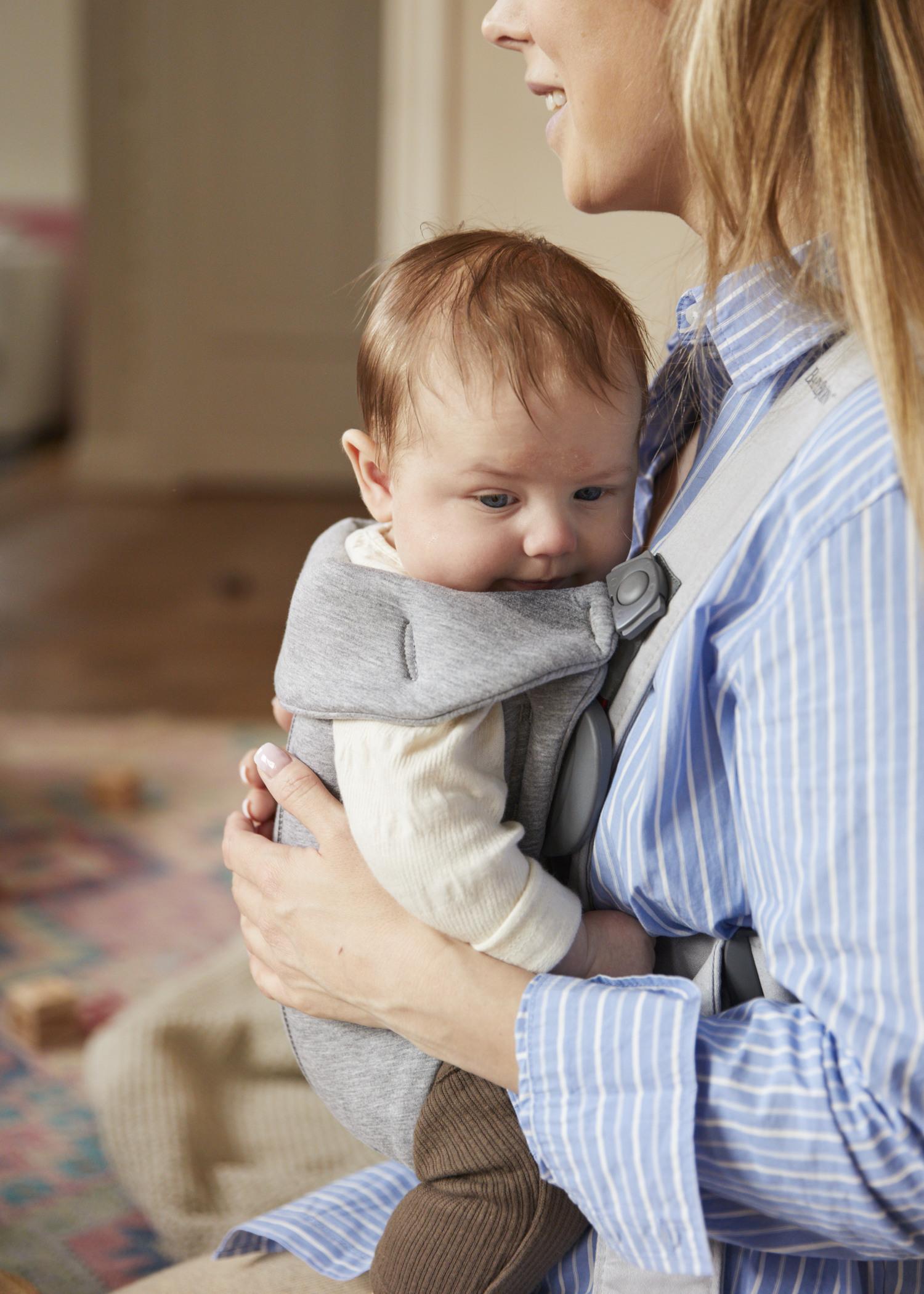 A woman in a blue striped shirt holds a baby wearing a gray carrier. The baby looks content and relaxed.