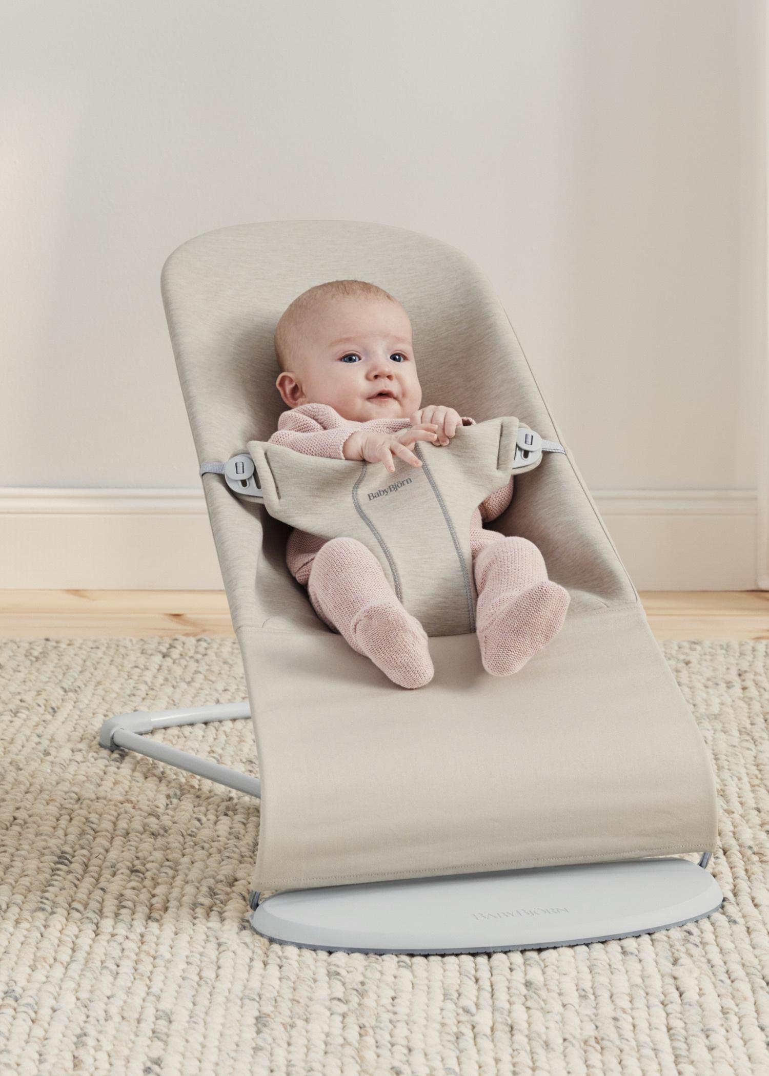 A baby in a pink outfit sits in a beige baby bouncer on a light-colored carpet.