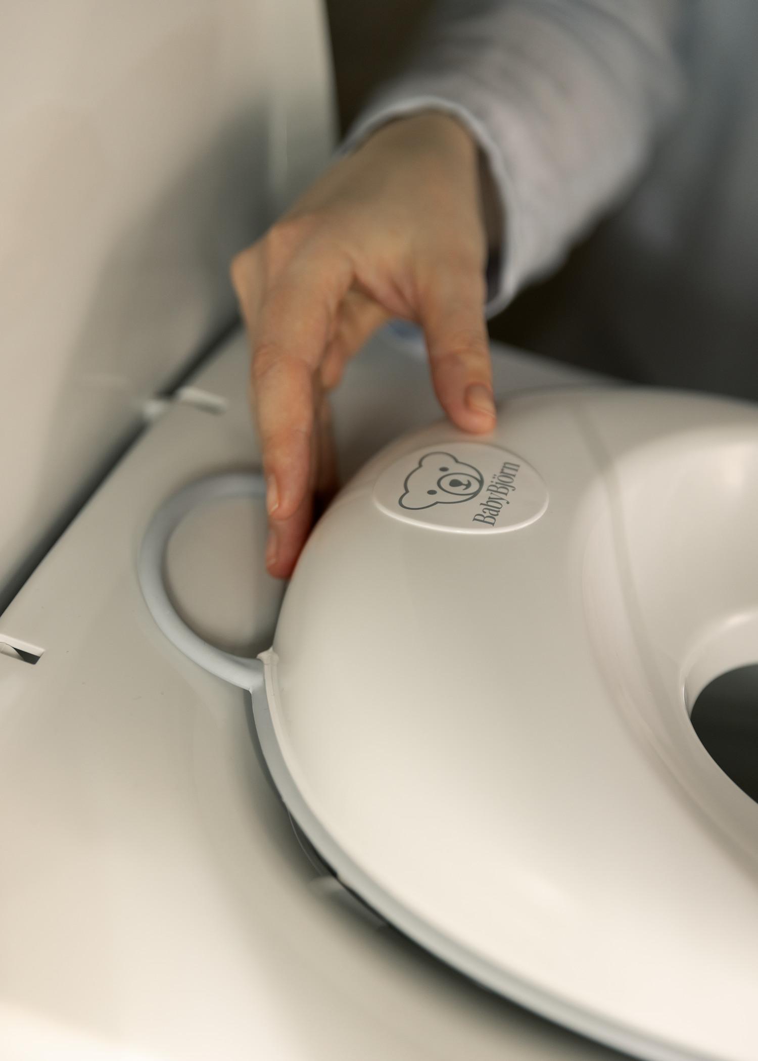 A person’s hand adjusting a white baby potty seat with a bear logo, attached to a toilet.
