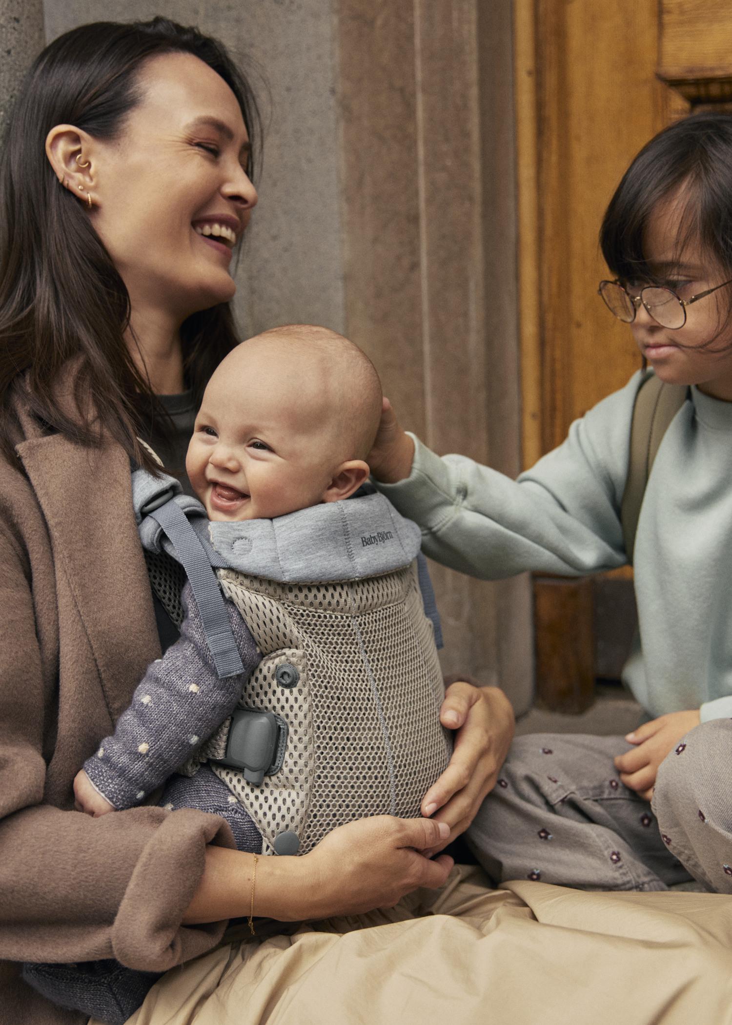 A smiling woman holds a happy baby in a carrier, while a child with glasses gently touches the baby's head. They sit together outdoors.