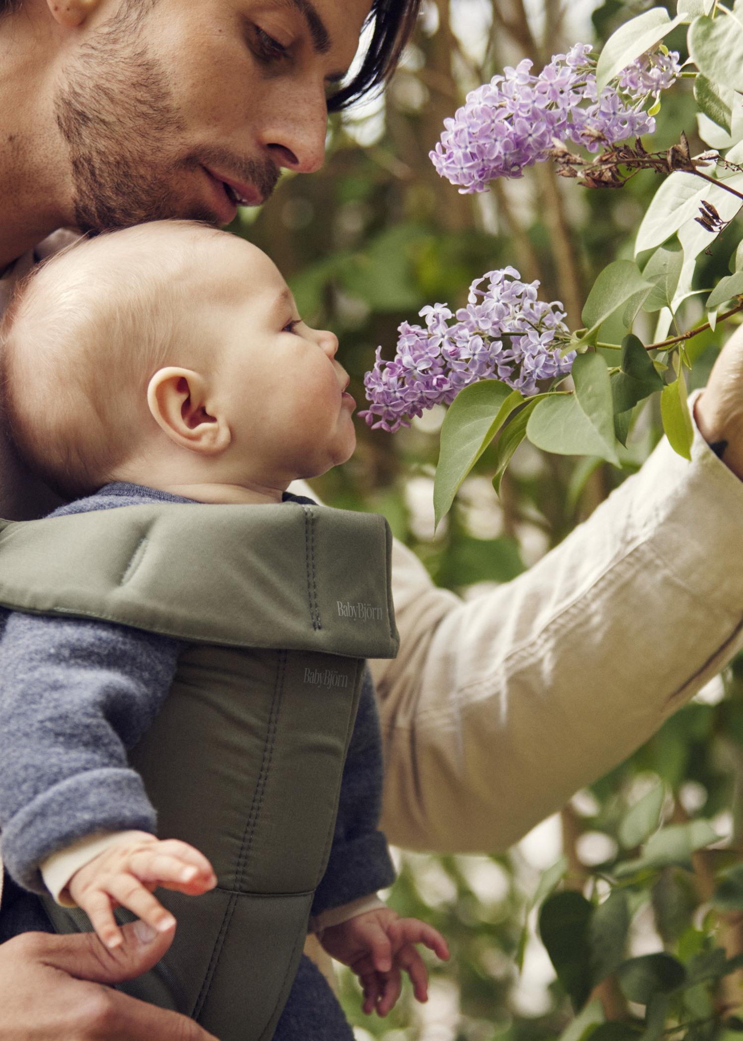 A man holding a baby in a carrier shows them purple flowers on a leafy branch. The baby leans forward, appearing to smell the blossoms.