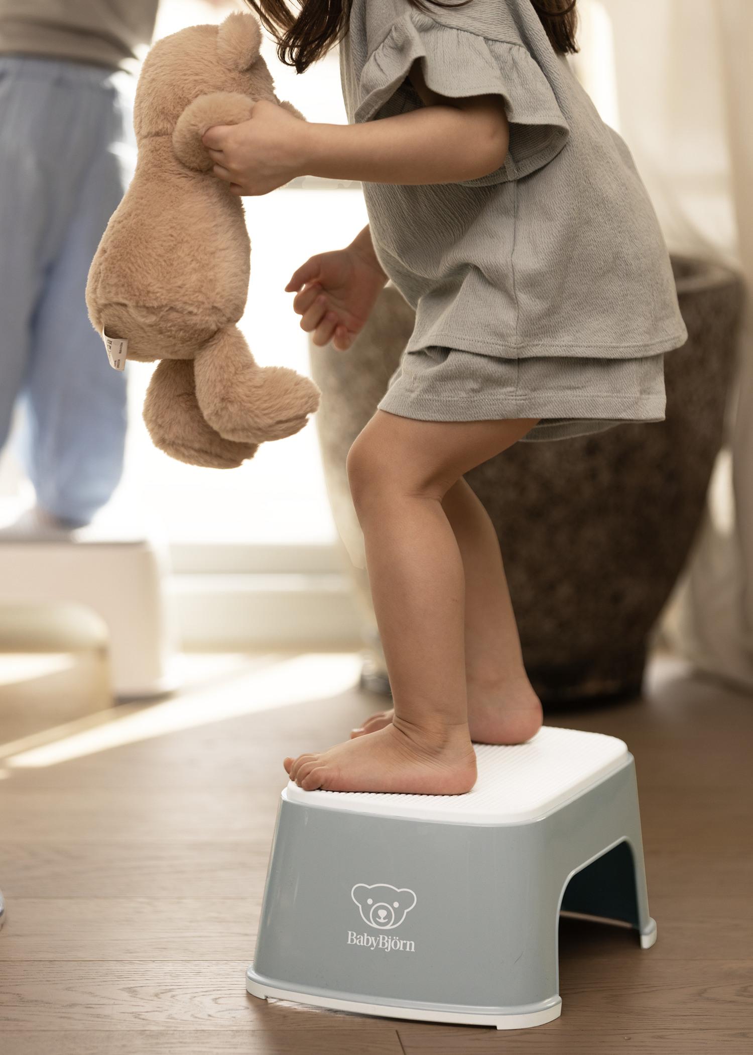 Child standing on a small stool, holding a teddy bear in a sunlit room.