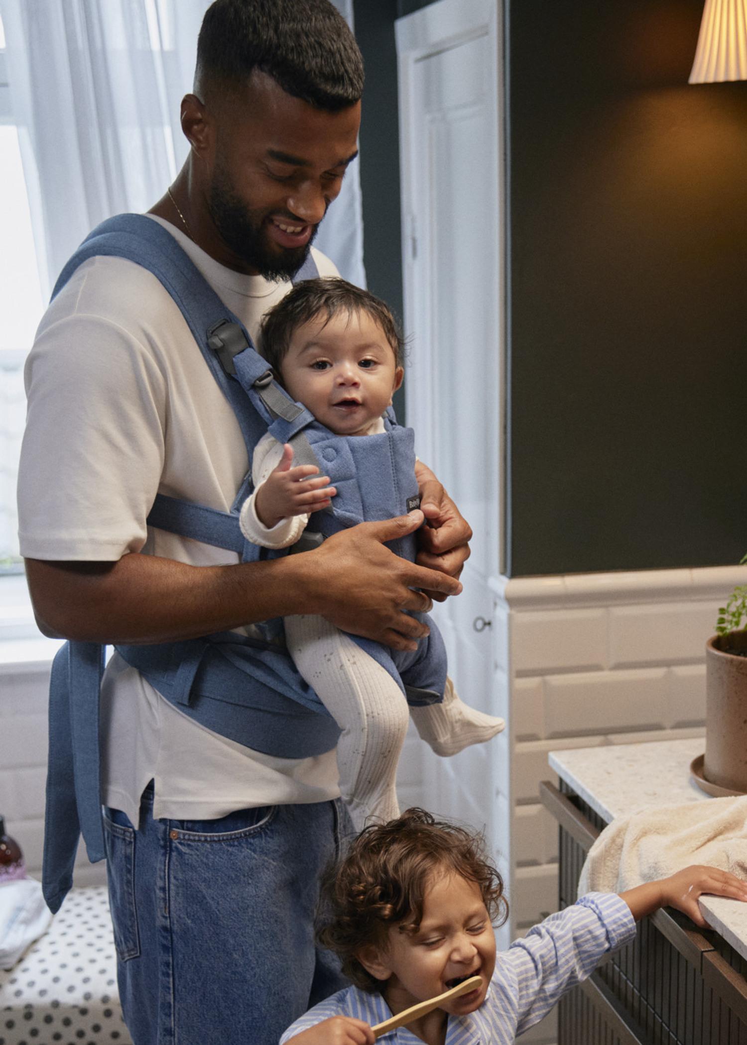 Father wearing a baby carrier holds an infant while a toddler brushes teeth at the sink in a cozy kitchen.