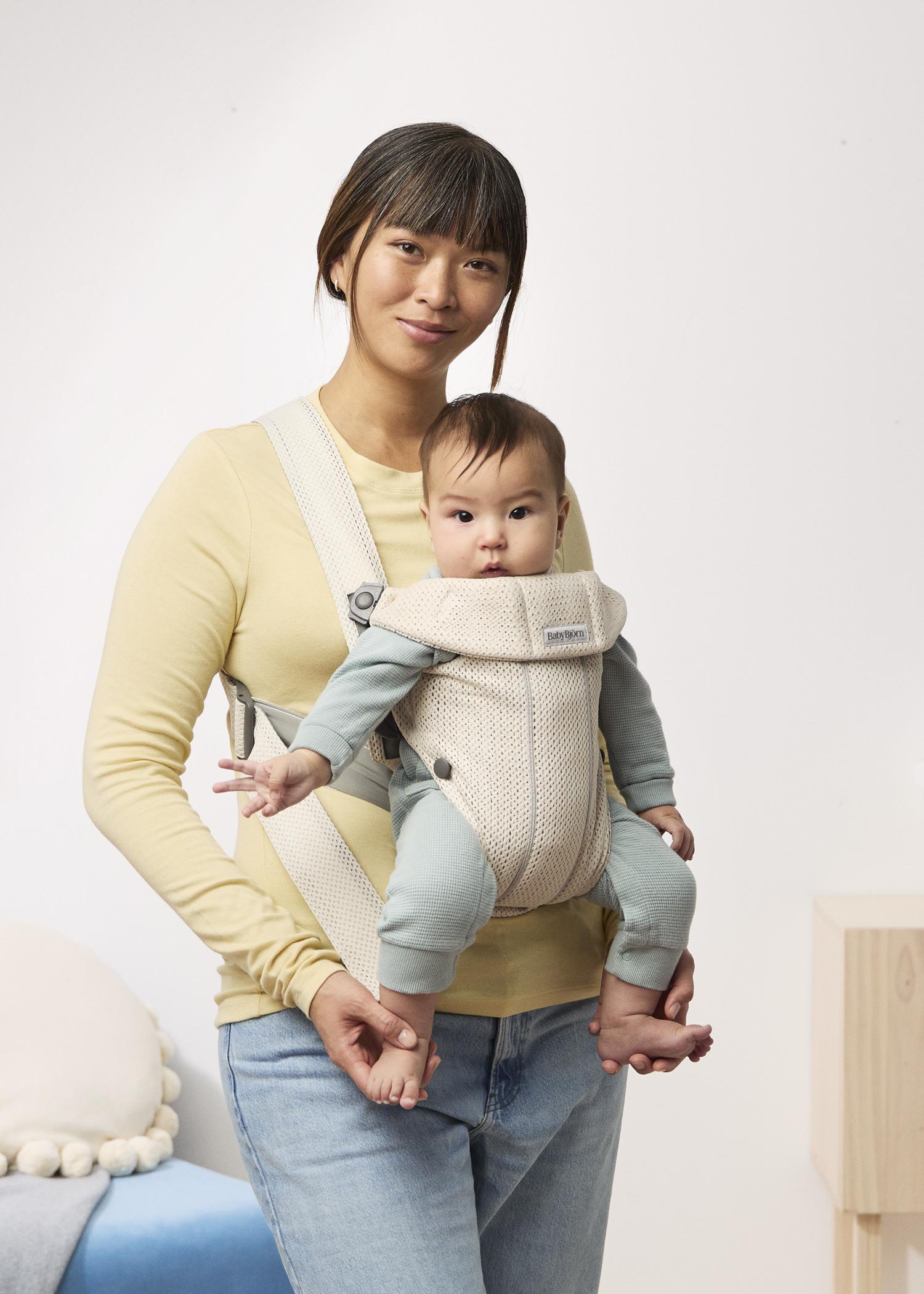 아기가 아기띠에 앉아 있고, 한 여성이 아기띠를 착용한 채 미소 짓고 있다. 배경은 심플한 실내이다.