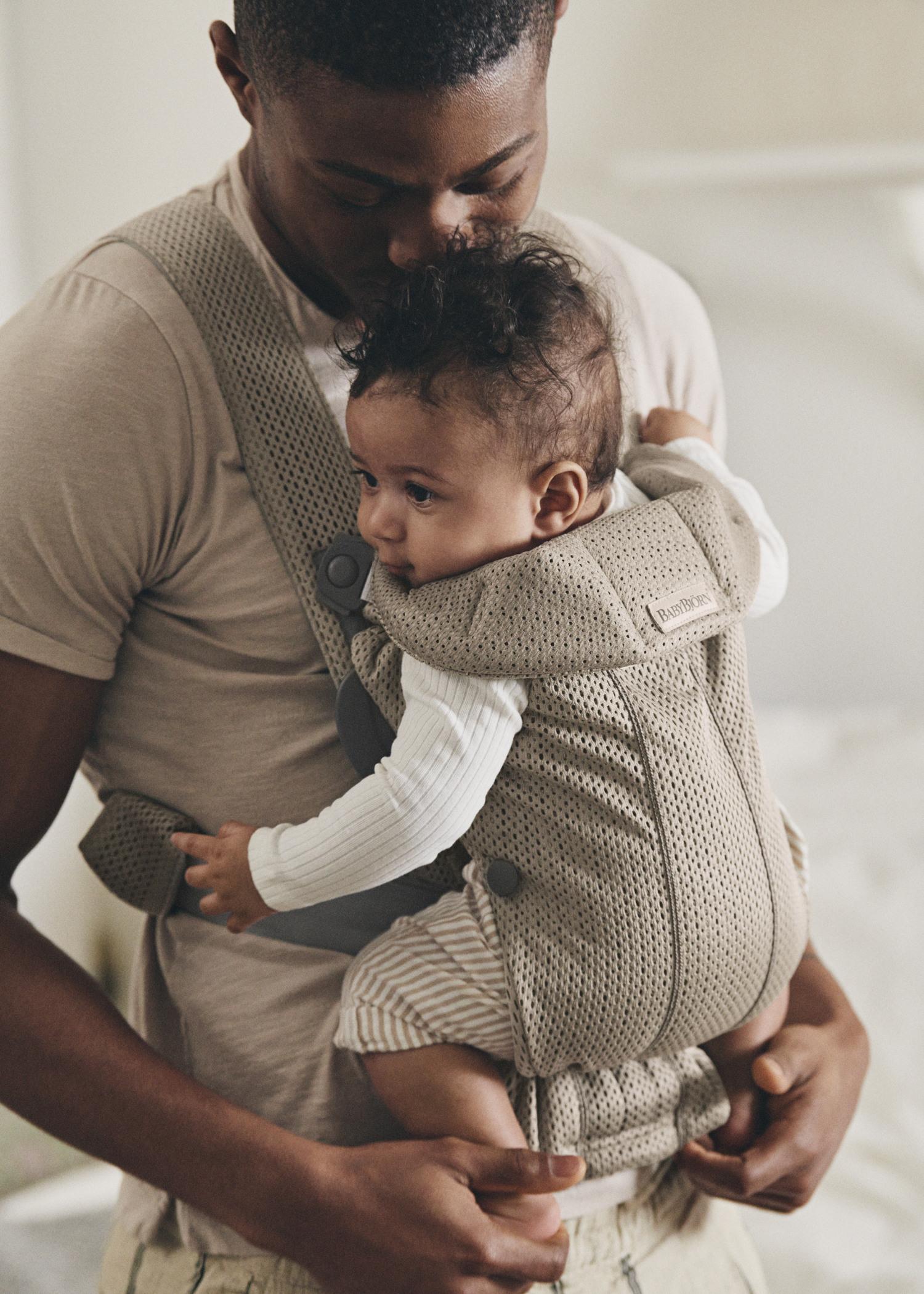 A man gently holds a baby in a beige carrier. The baby is facing forward, wearing a white shirt and striped pants. The setting is soft and cozy.