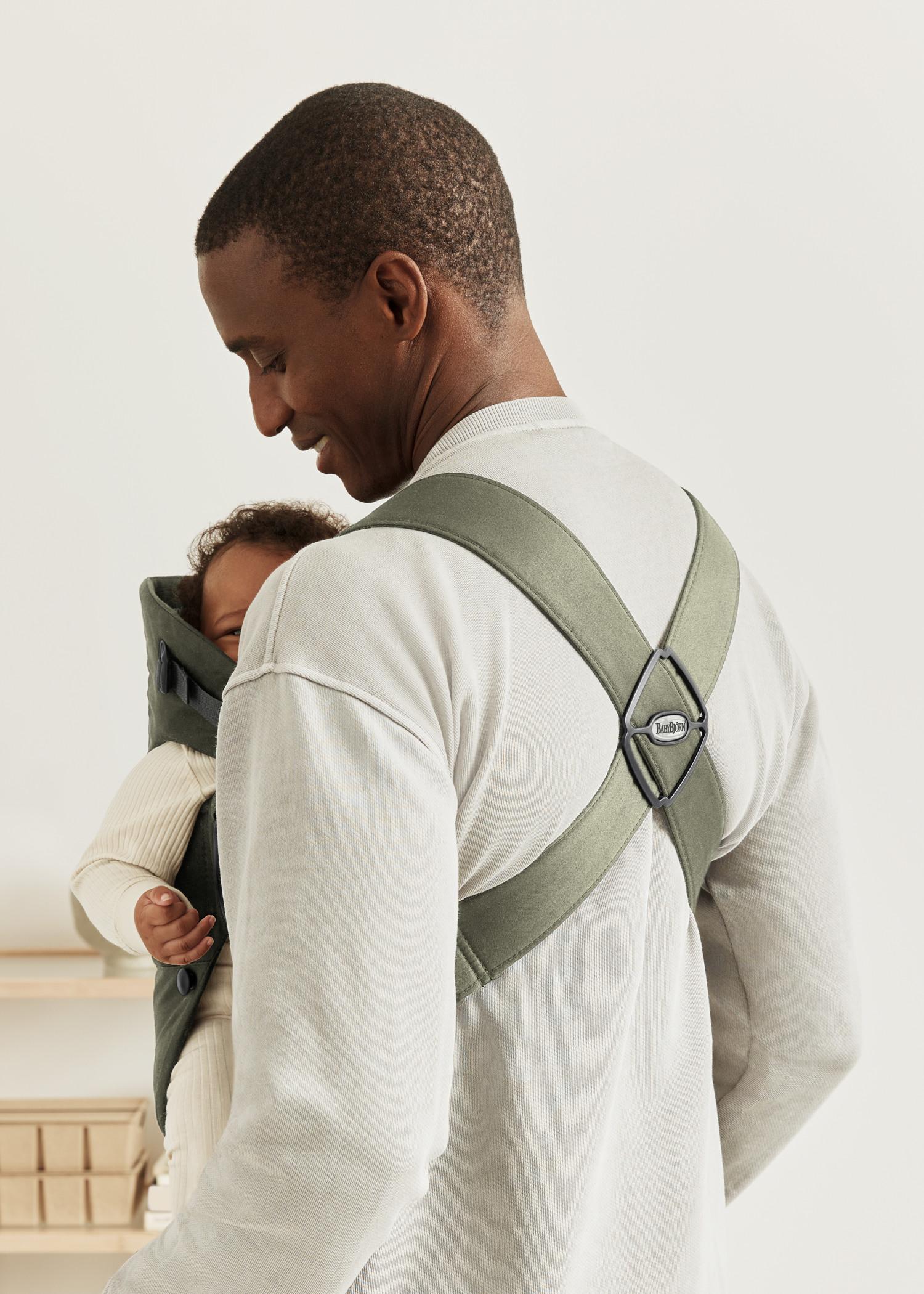 A person in a light sweater carries a baby in a green baby carrier with crossed straps, facing forward and smiling gently.