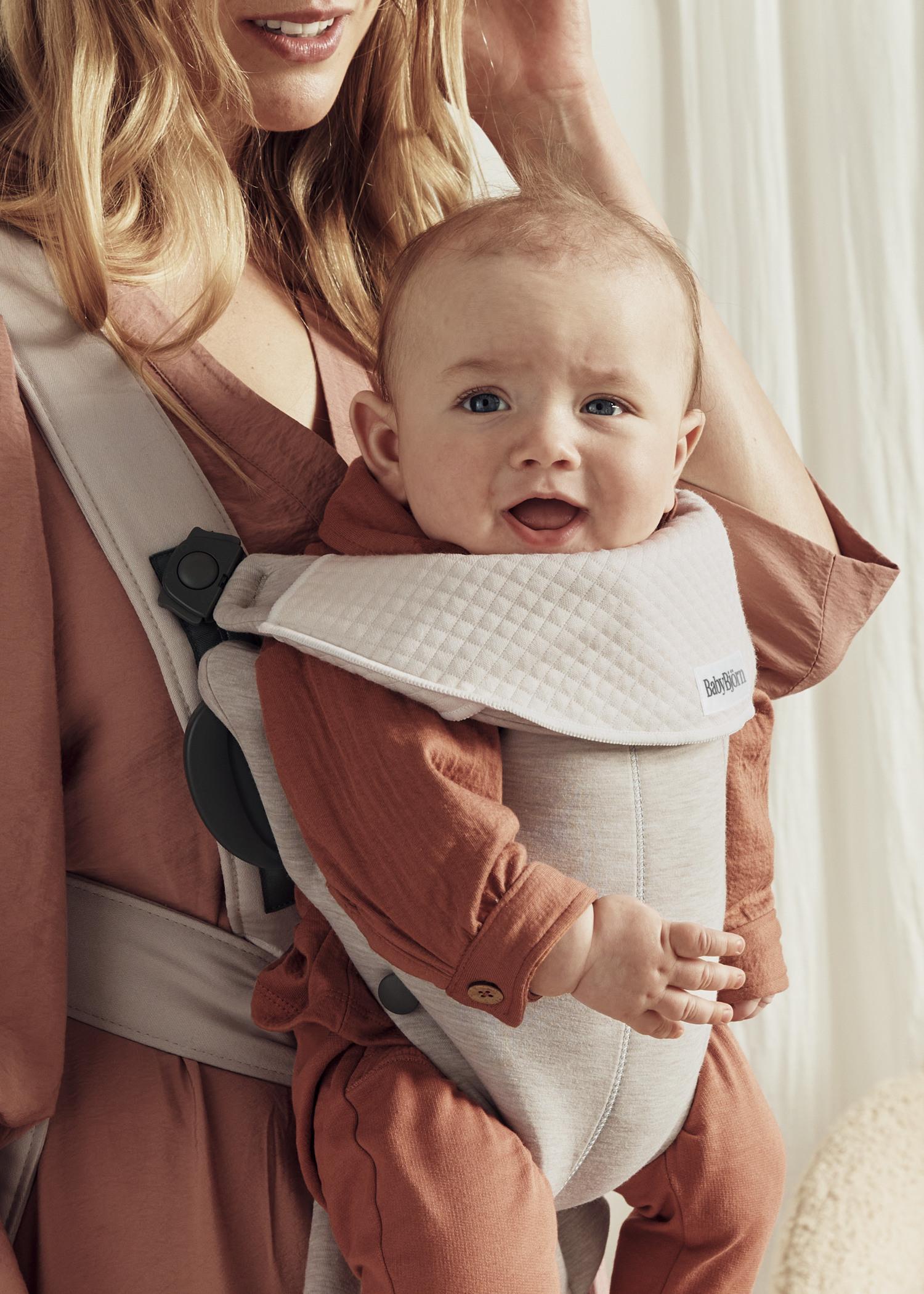 A smiling baby in an orange outfit is carried in a soft, beige baby carrier. The carrier is worn by a woman with long, blonde hair.