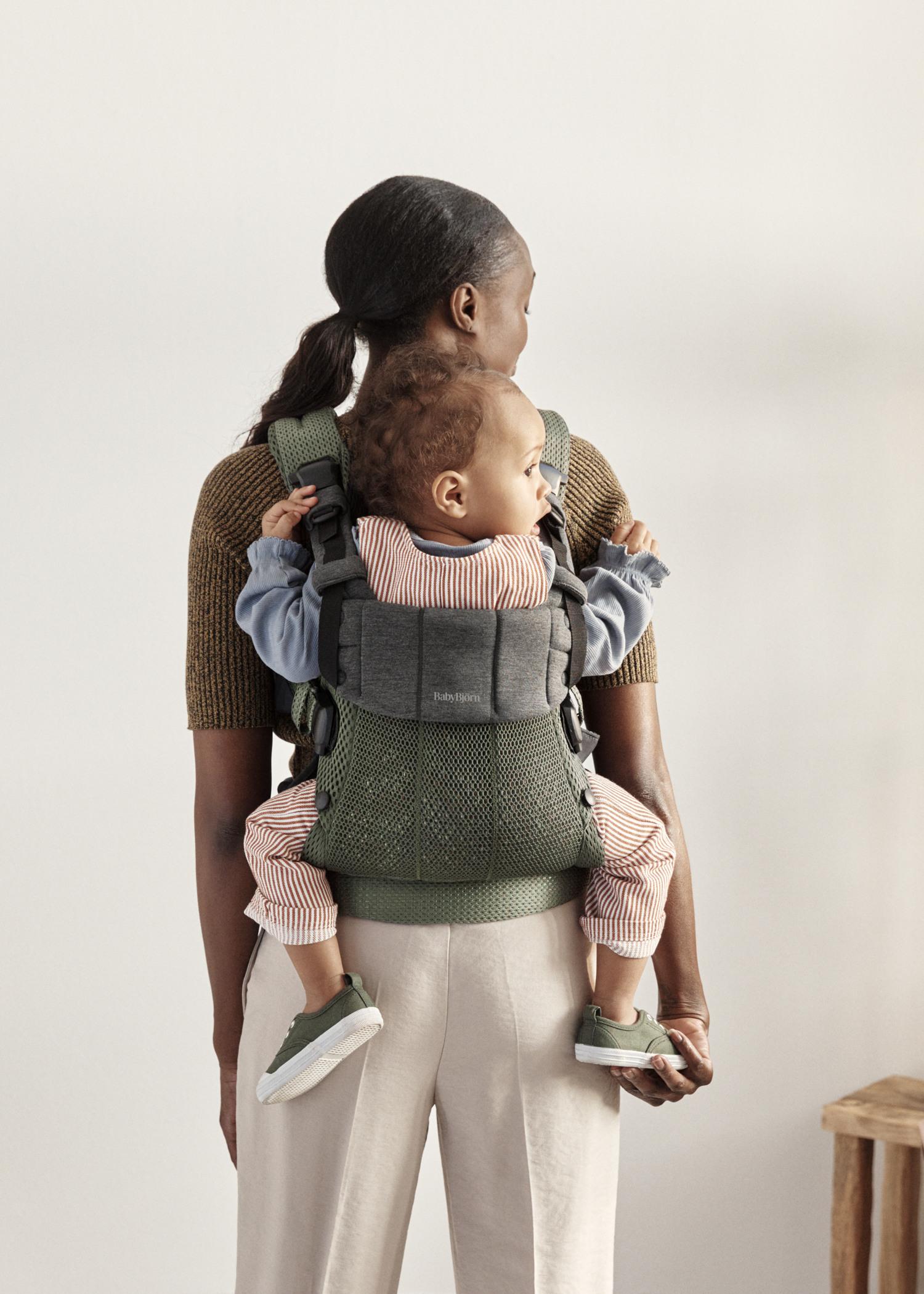 Woman with hair in a ponytail, wearing a brown top and beige pants, carries a baby in a green and gray carrier on her back.