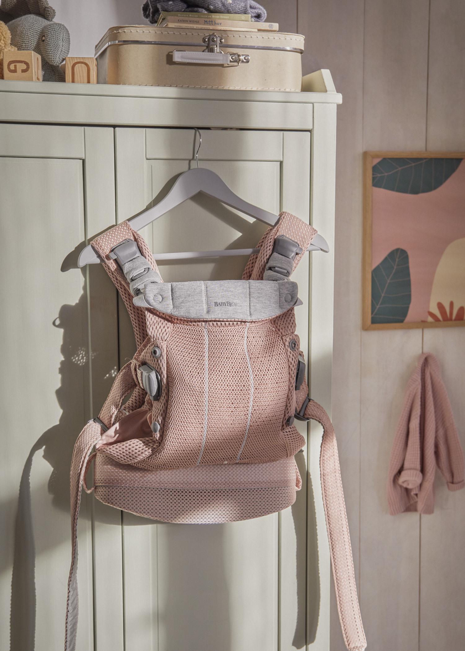A pink baby carrier hangs on a green wardrobe door, with a suitcase on top and a colorful wall art in the background.