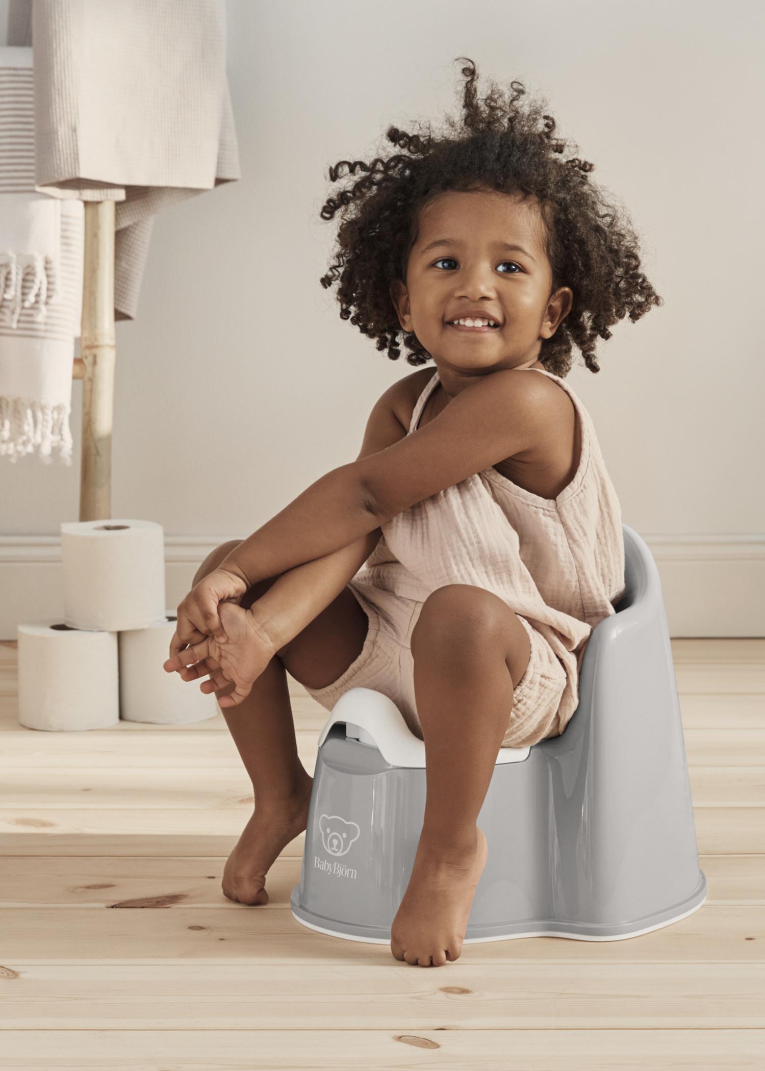 A child with curly hair sits on a gray potty chair, smiling. The room has wooden floors, rolled-up towels, and toilet paper in the background.