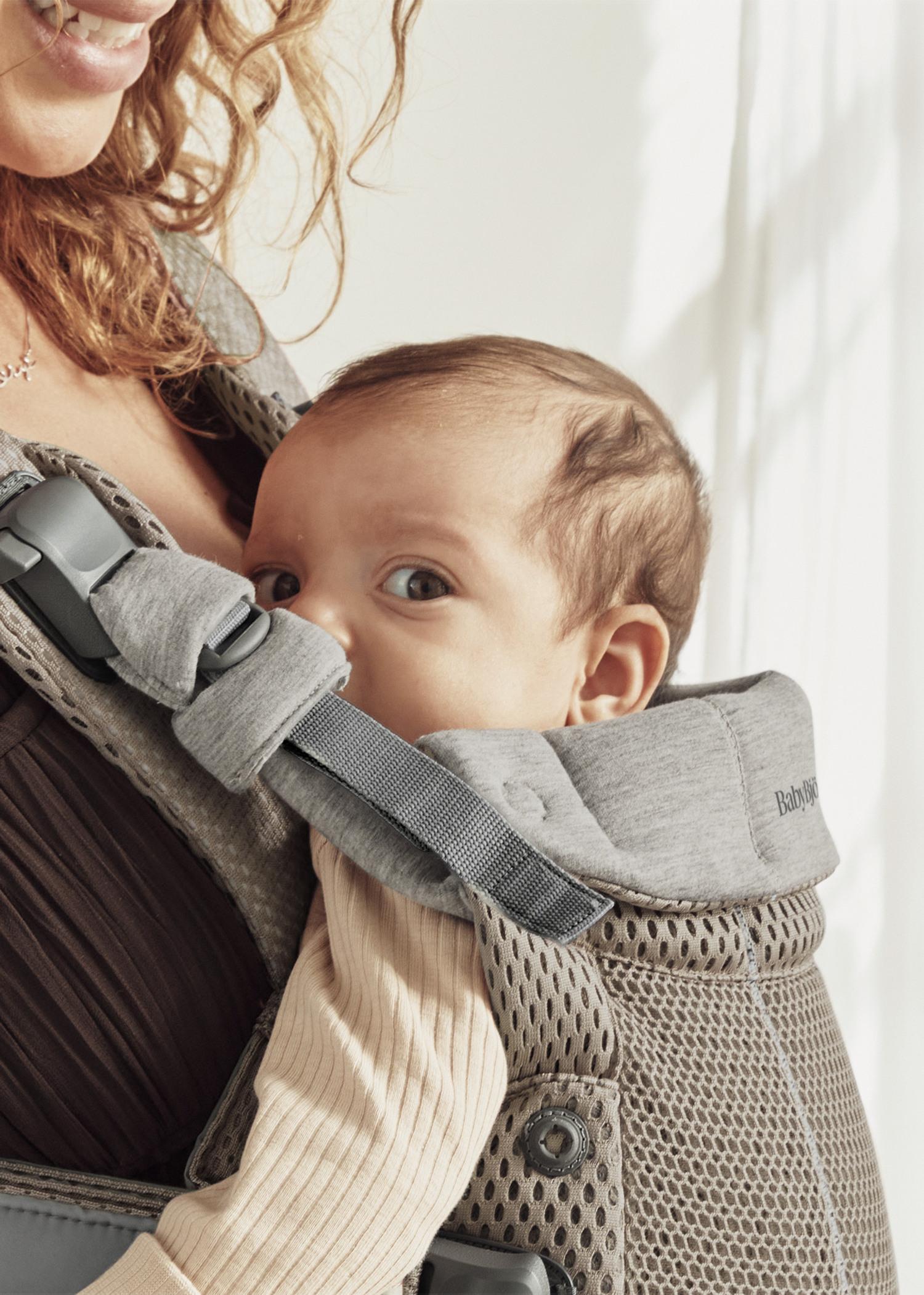 A baby peeks out from a gray beige baby carrier, held by a woman with curly hair, against a softly lit background.