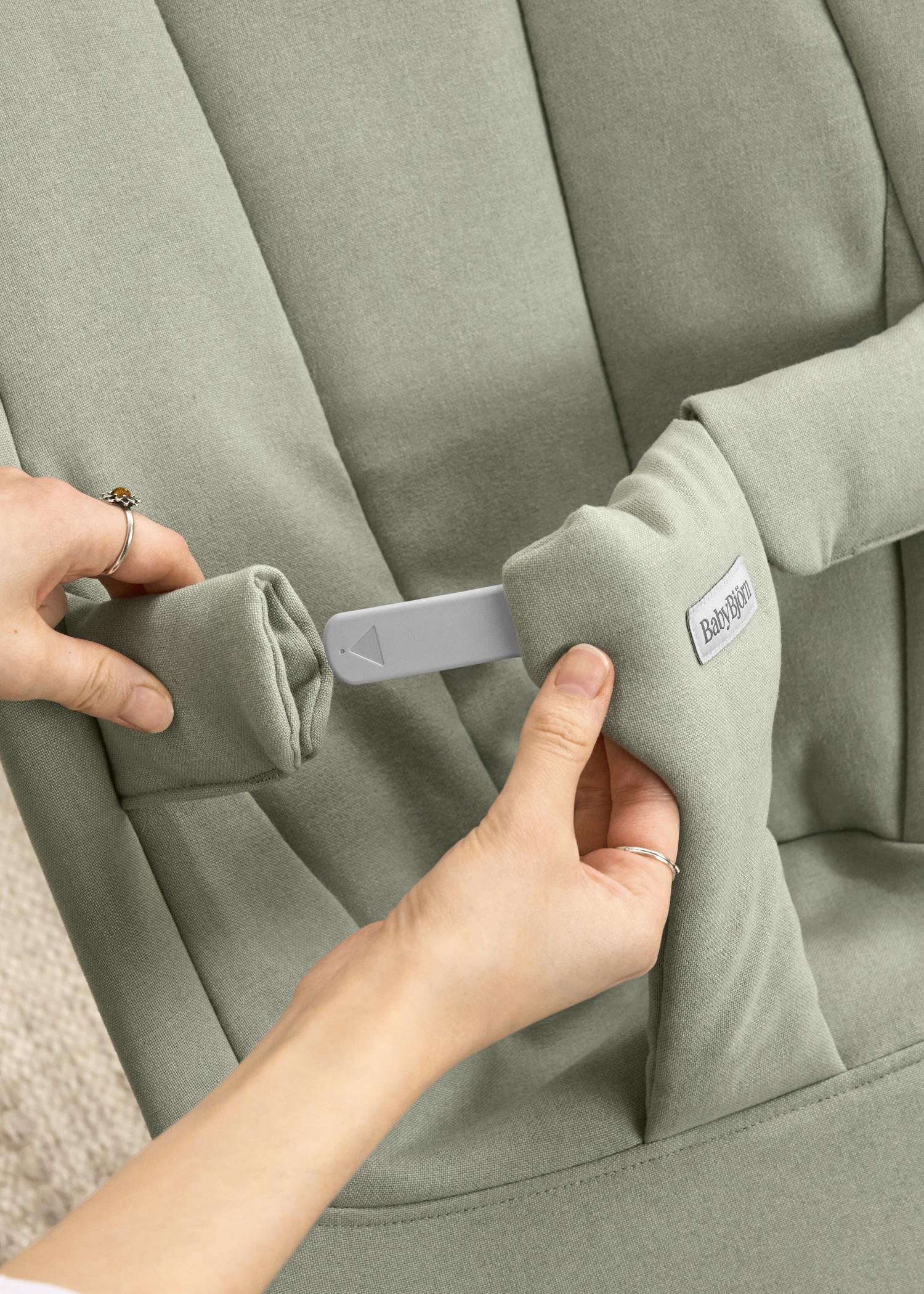 Hands adjusting a light green baby bouncer with a BabyBjörn label, showcasing a strap and padded cover.