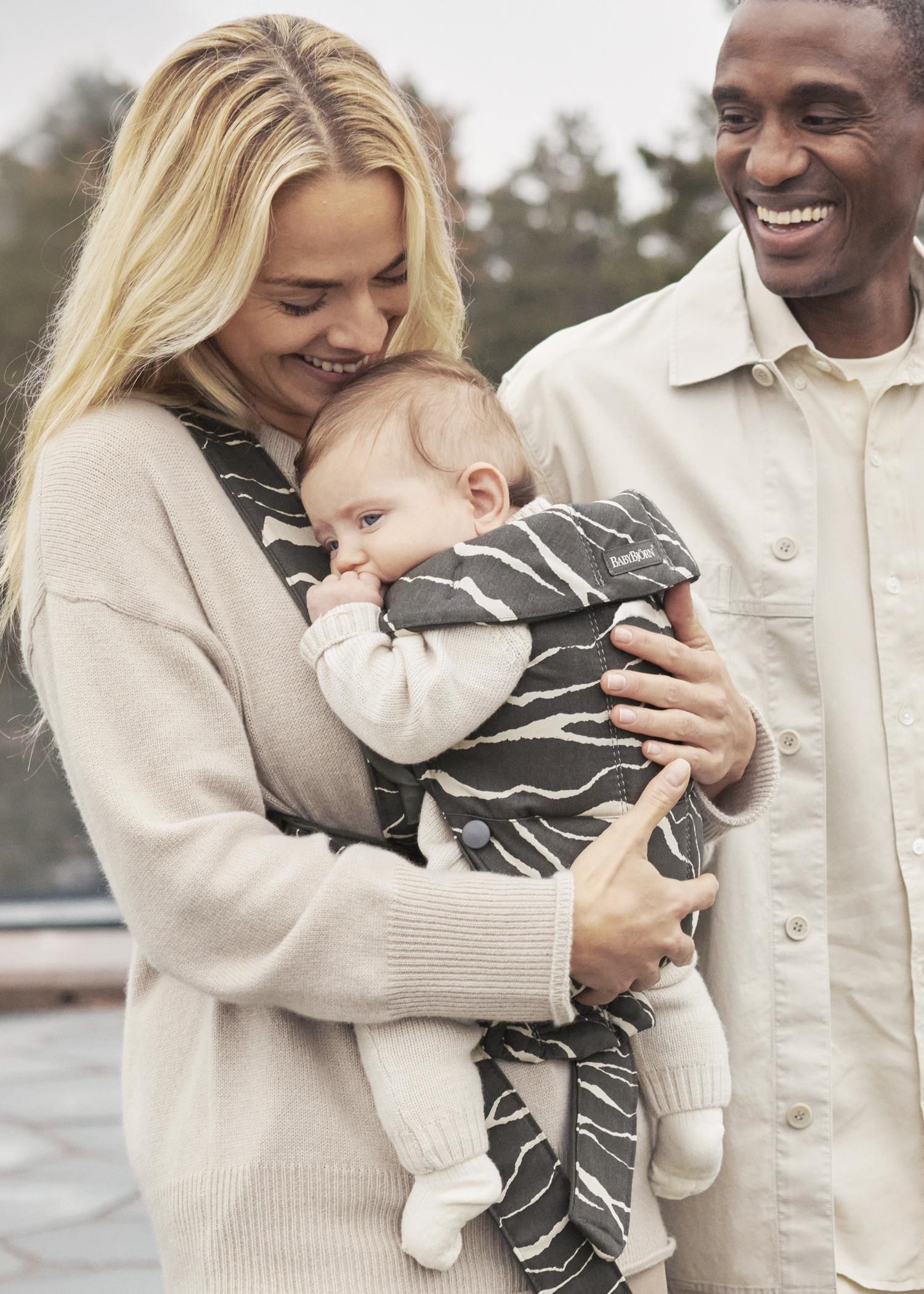 A smiling couple holds a baby in a patterned carrier. The woman, with long blonde hair, embraces the baby, while the man smiles beside them.