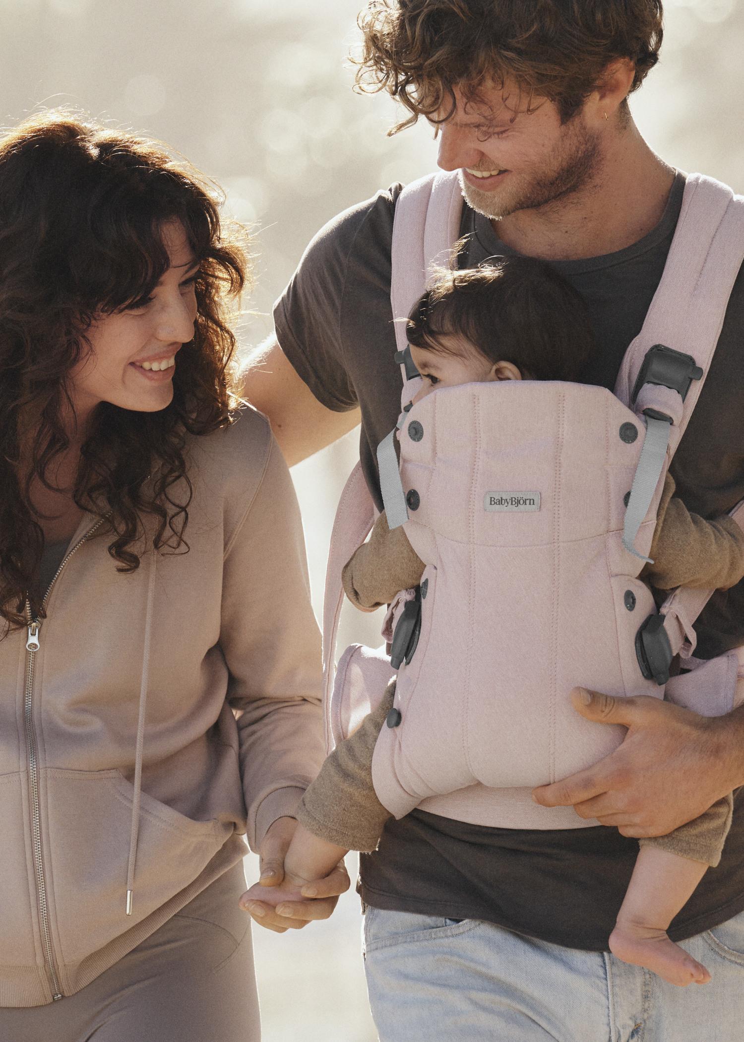 A couple walks outdoors, smiling at each other. The man carries a baby in a pink carrier on his chest, and the woman holds the baby's hand.