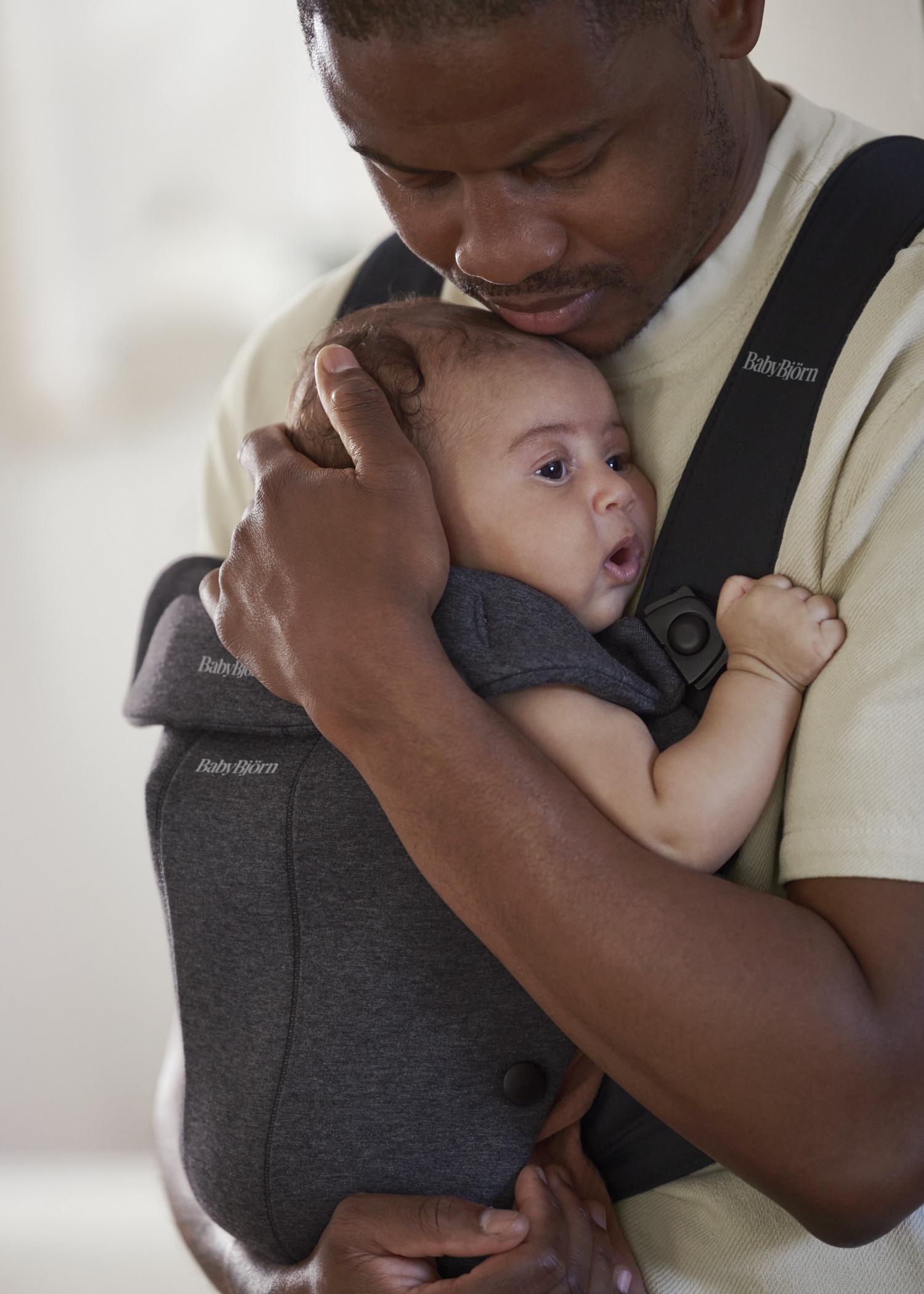 A man tenderly holds a baby in a BabyBjörn carrier, gently resting his head on the baby's.