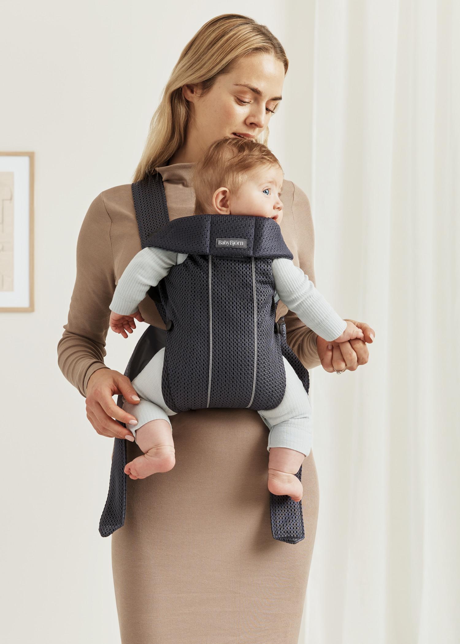 A woman stands with a baby in a front-facing baby carrier. The baby is wearing light-colored clothing, and the woman is holding the baby's hand.