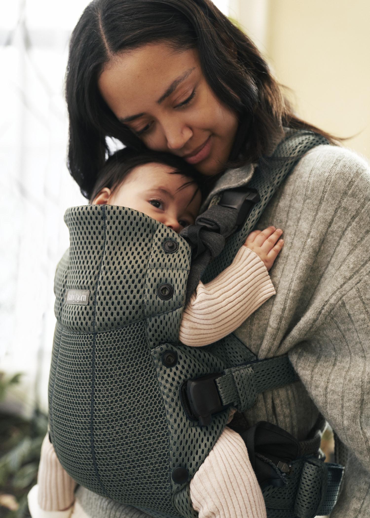 A woman smiles gently, cradling a baby in a green mesh baby carrier, both wearing cozy sweaters in a softly lit room.