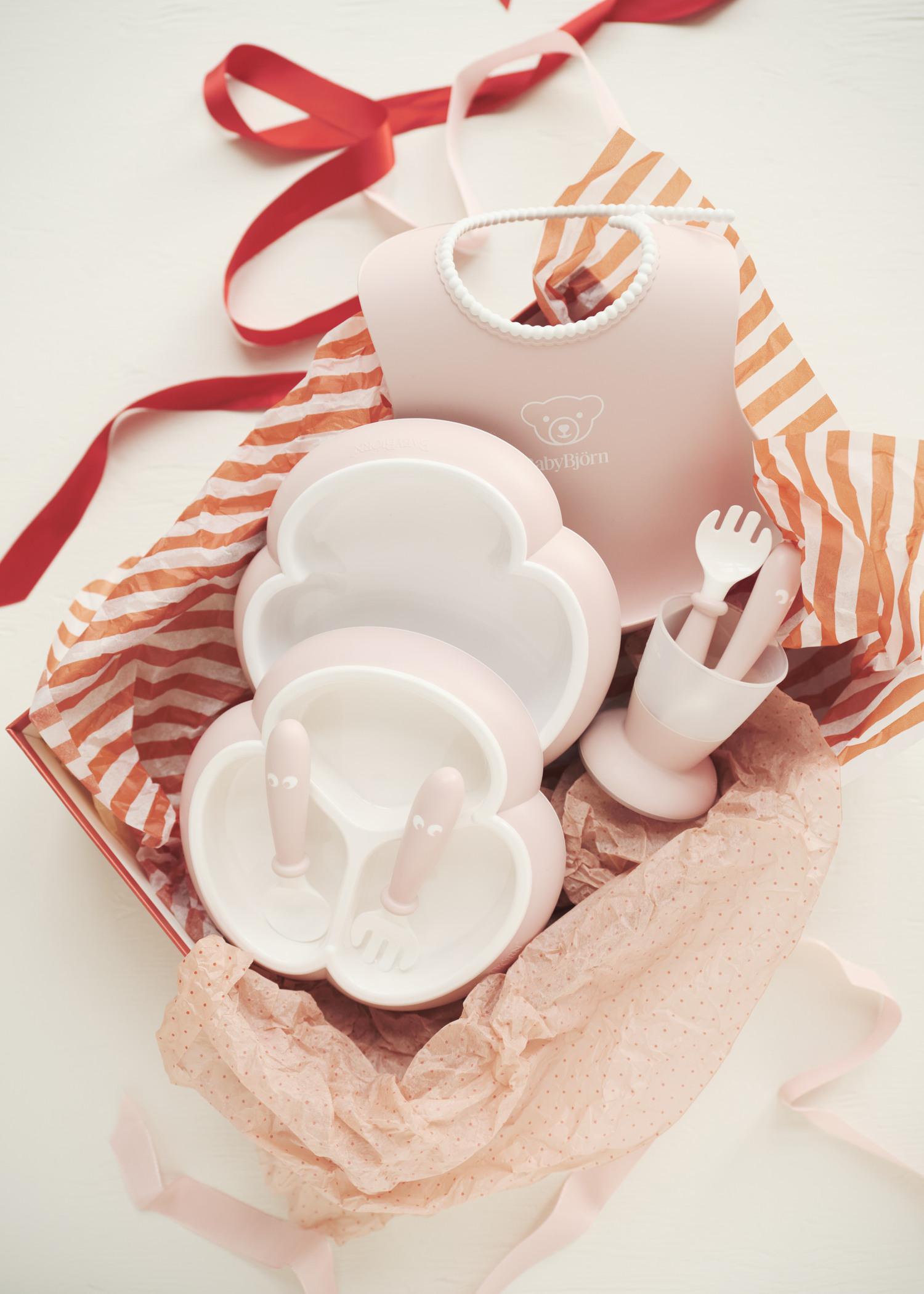 Pink and white baby dining set with plates, utensils, and a bib in a gift box with red and pink tissue paper and ribbon.