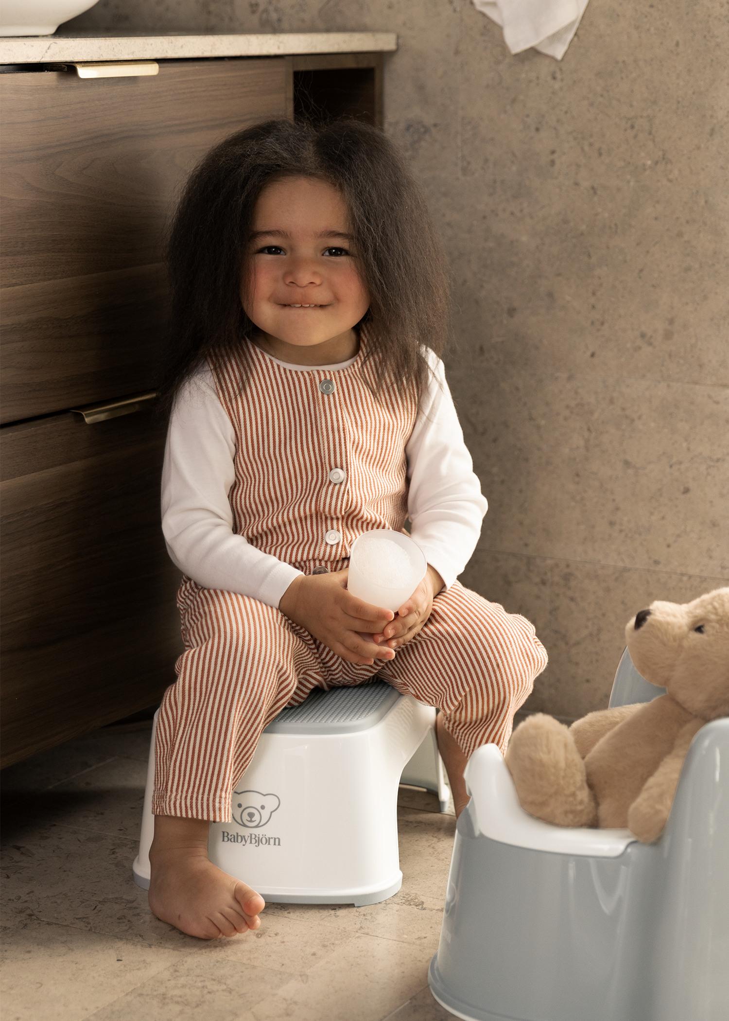 Child with long hair in striped overalls sits on a step stool, holding an egg, near a toy bear on a potty.