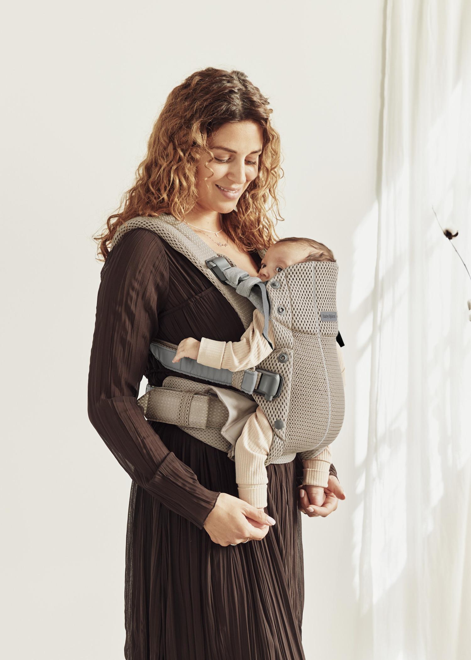 A woman with curly hair smiles while holding a baby in a beige baby carrier. She is standing in a softly lit room with a white curtain.