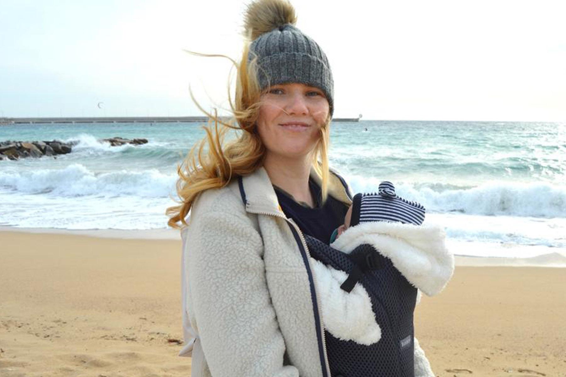 Smiling woman in a knit hat and fleece jacket carrying a bundled baby in a carrier on a windy beach.